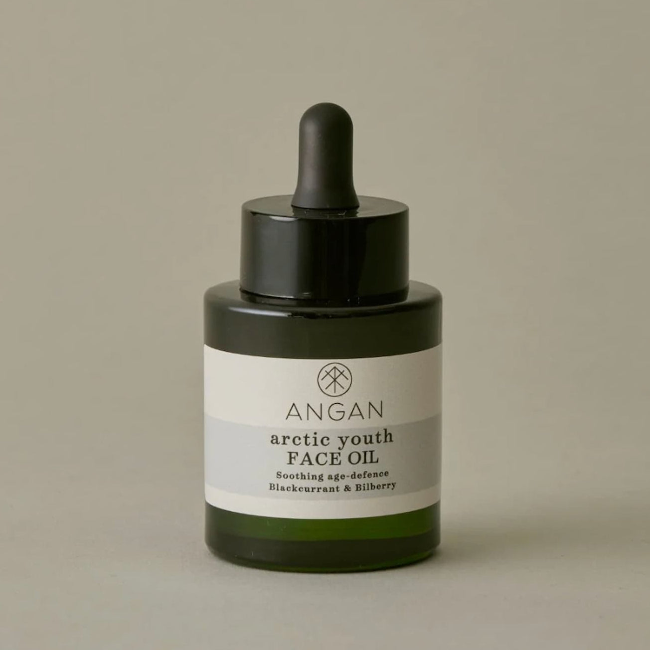 Angan Arctic Youth Face Oil in a dark green glass dropper bottle with a black dropper cap and a white label, set against a warm gray background.