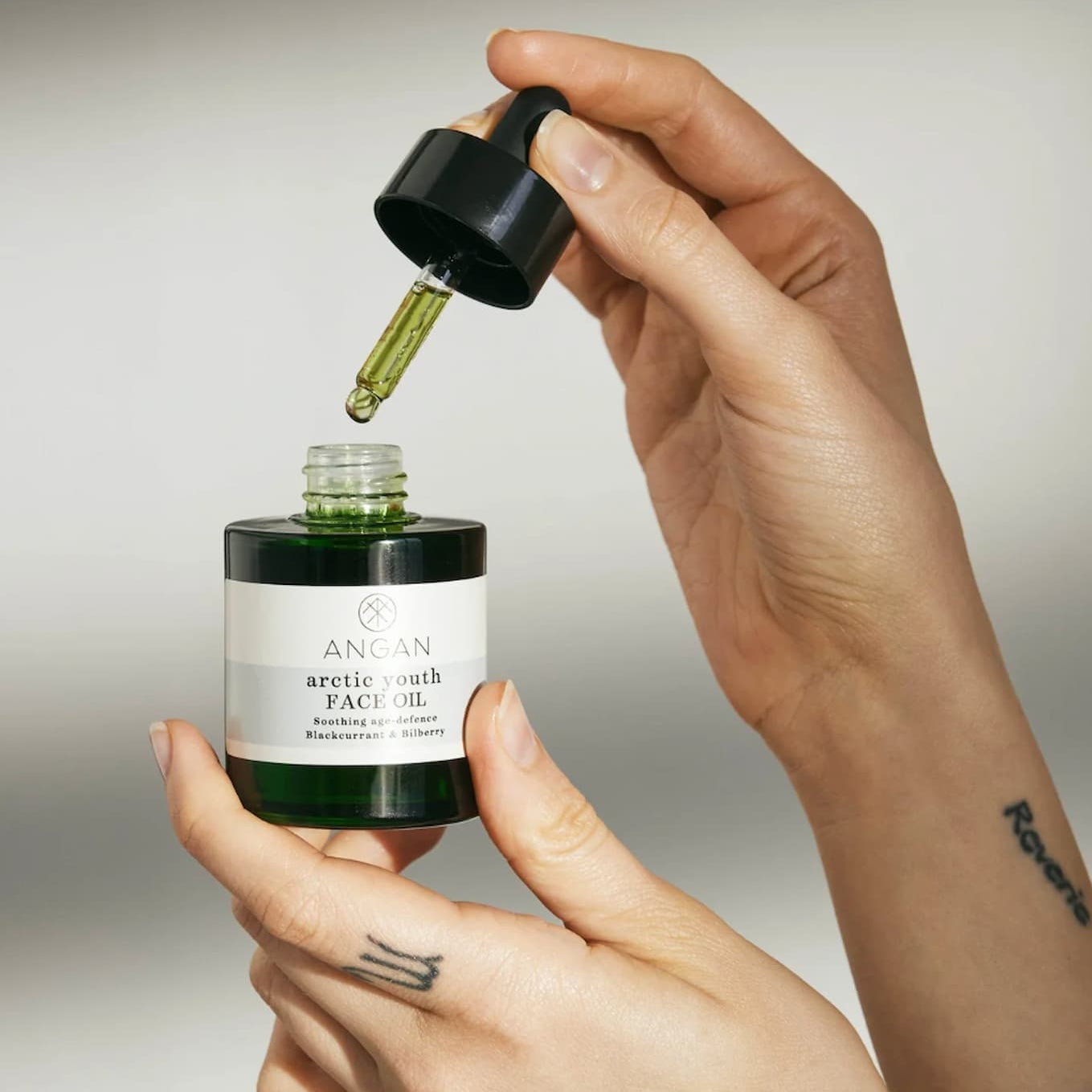 A close-up of a person's hands holding a bottle of Angan Arctic Youth Face Oil, with the black dropper cap lifted to release a drop of green oil above the open bottle against a light gray background.