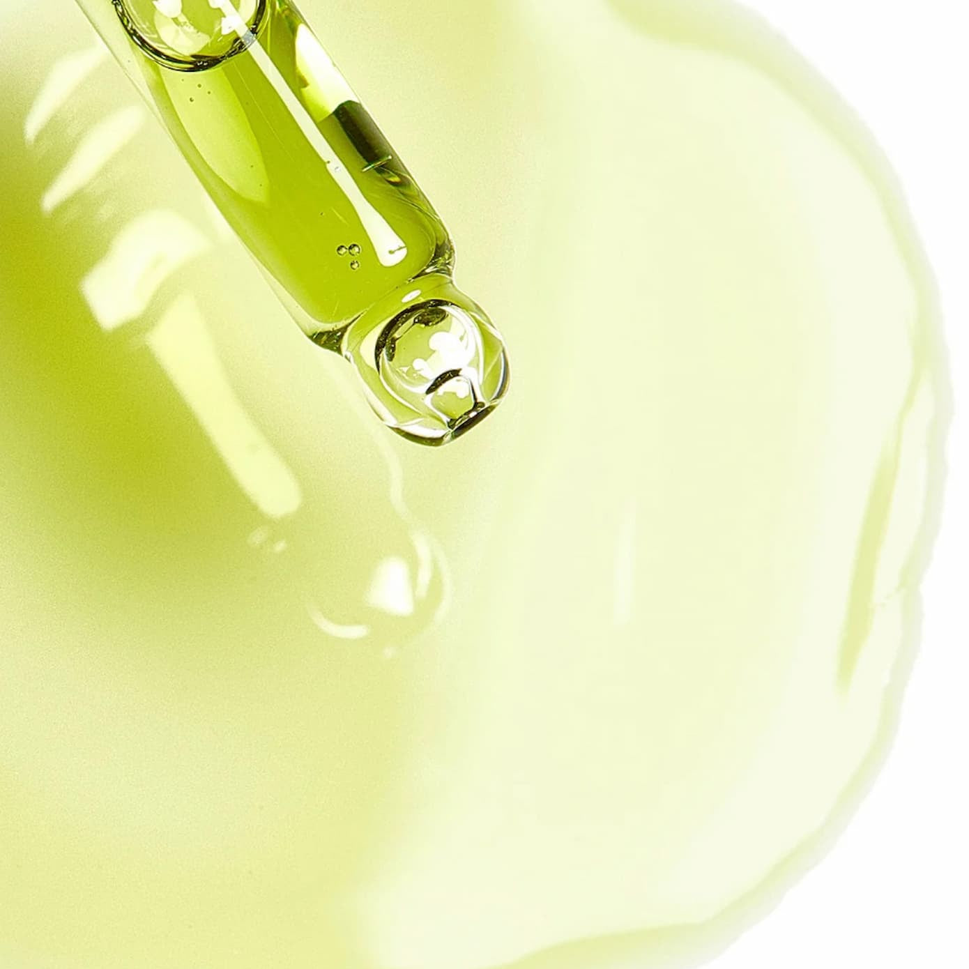 A close-up of a clear glass dropper dispensing pale yellow-green face oil onto a white surface, with a glossy pool of the same translucent product below.