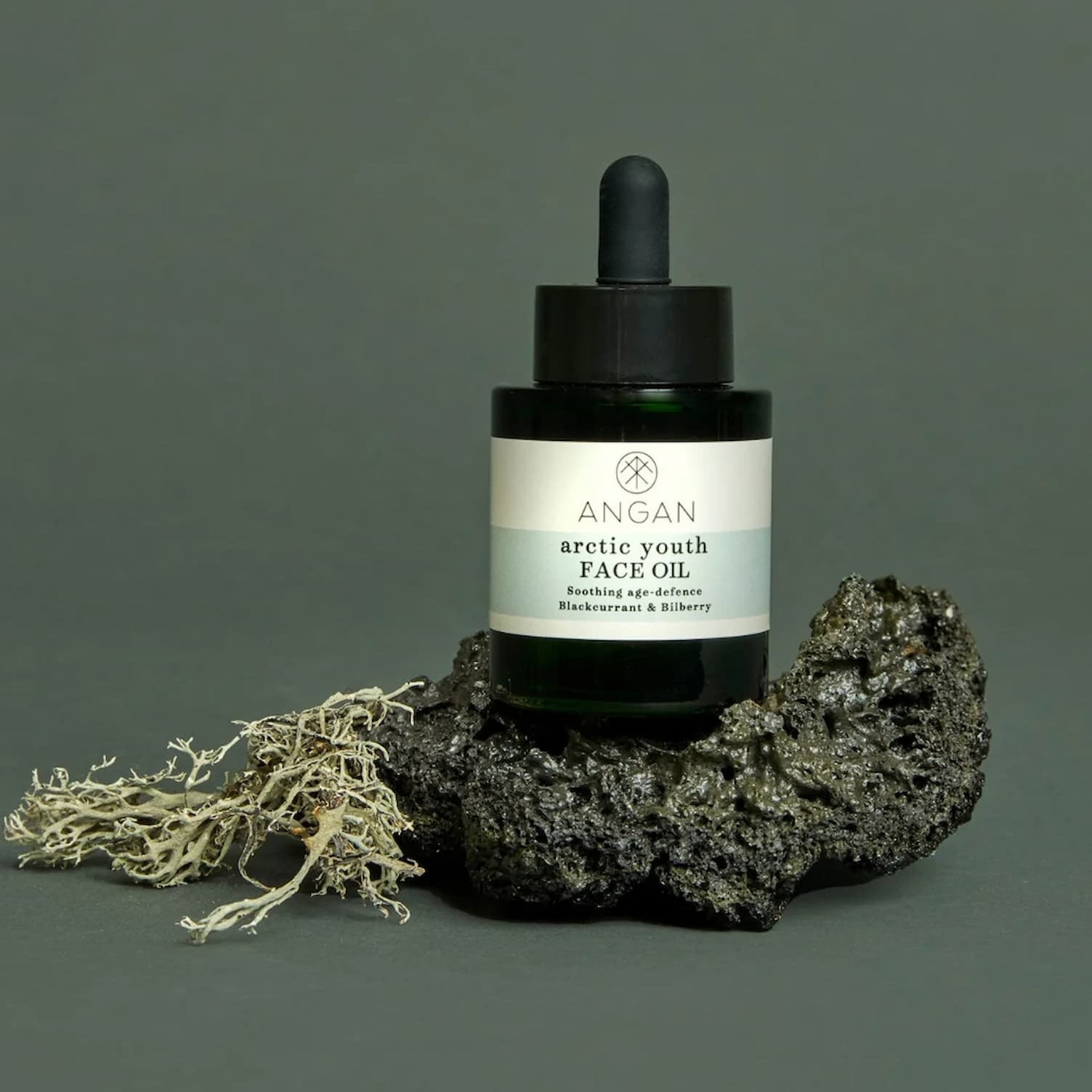 A dark green glass dropper bottle of ANGAN Arctic Youth Face Oil, styled on a textured dark rock with dried moss against a solid dark green-gray background.