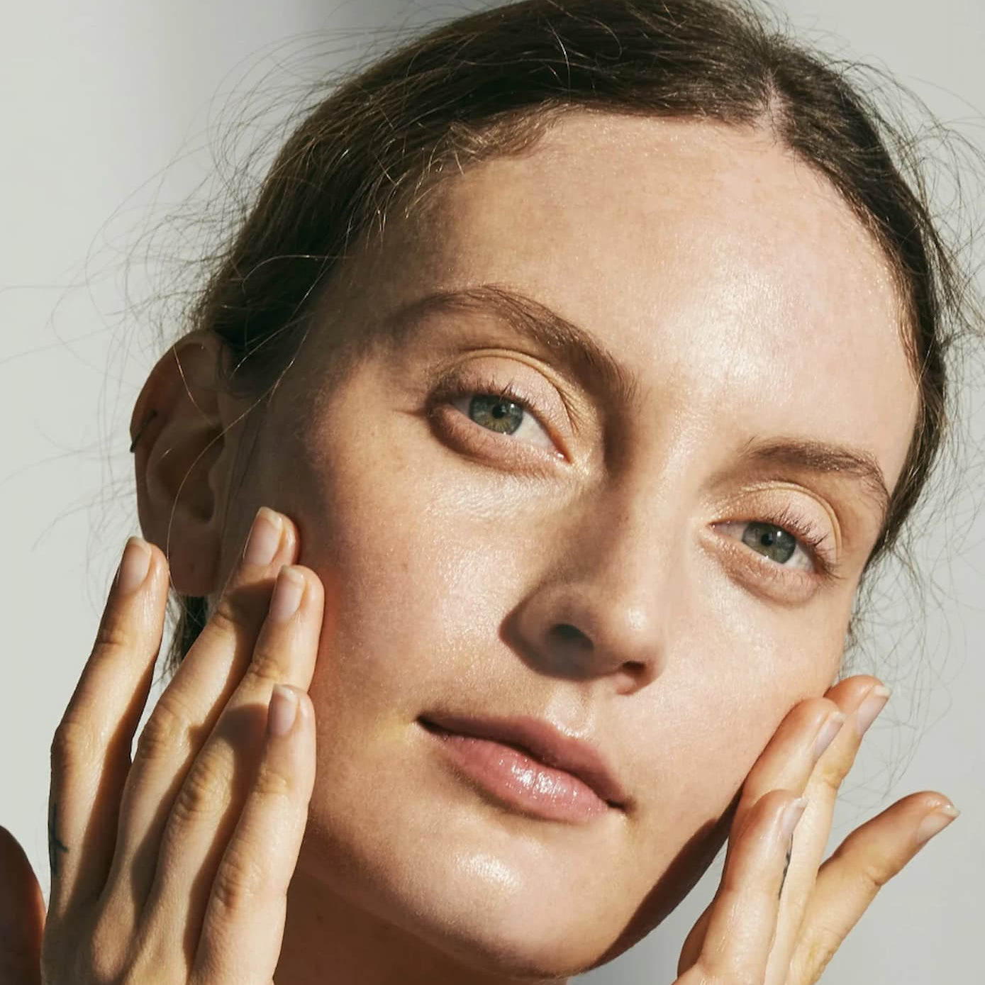 A close-up portrait of a woman with bare skin and her hands touching her cheeks, set against a plain light gray background.
