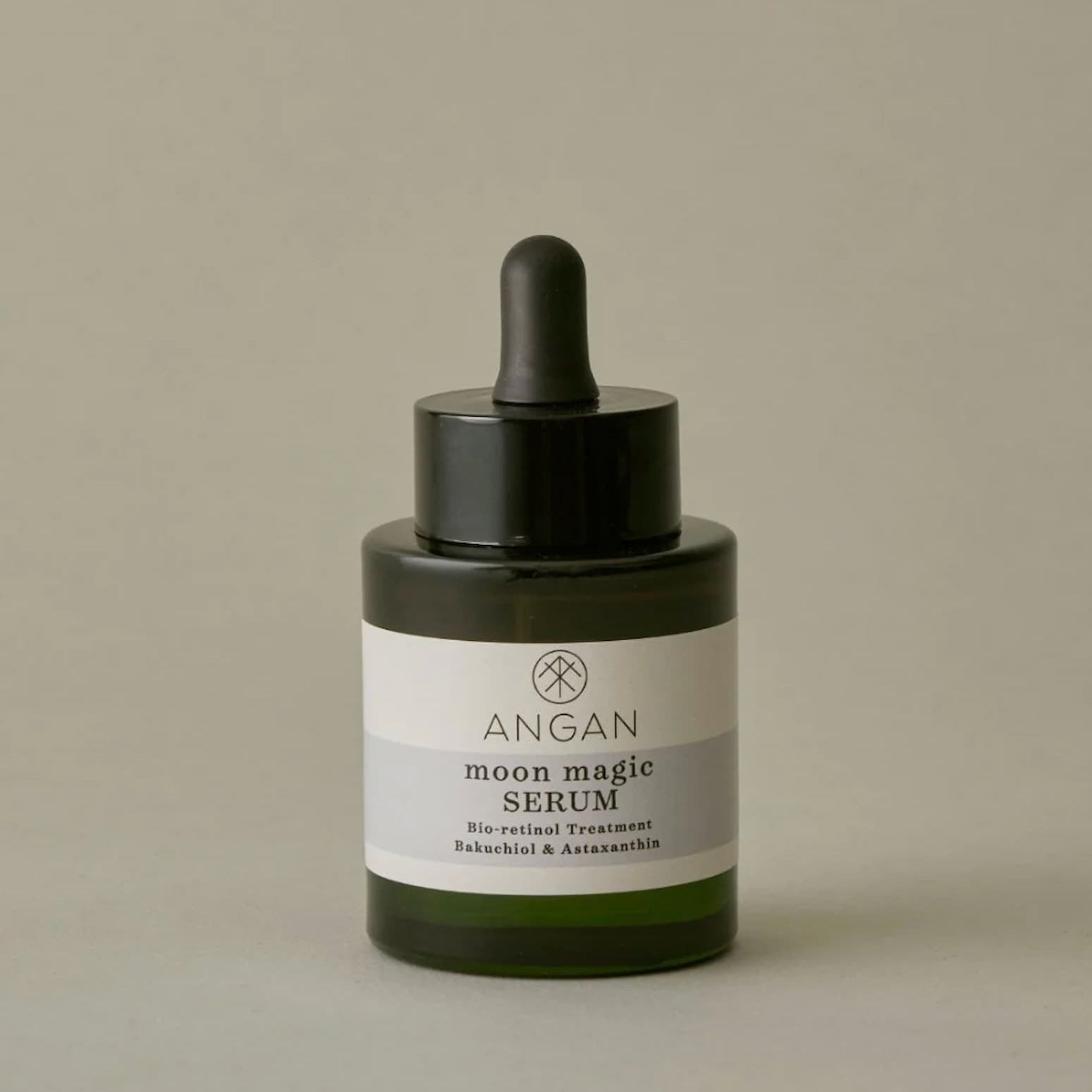 Angan Moon Magic Serum in a dark green glass dropper bottle with a black cap and a white label, set against a solid warm gray background.