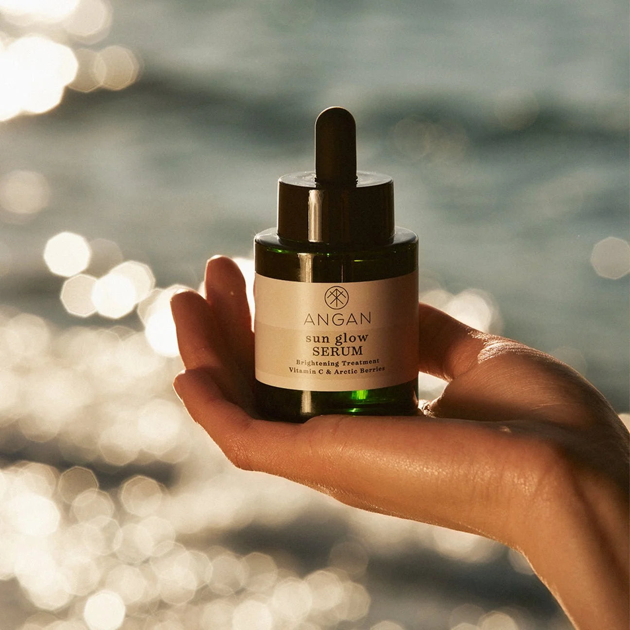 Hand holding a bottle of 'ANGAN sun glow SERUM' with a blurred natural background