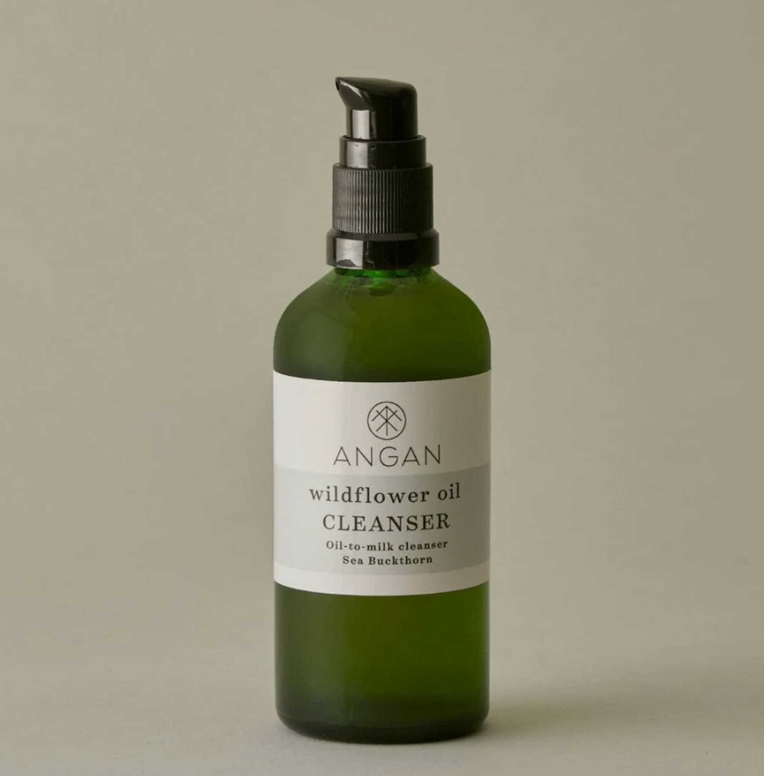 A green glass pump bottle of Angan Wildflower Oil Cleanser with a black pump dispenser and a white label, set against a solid neutral gray background.