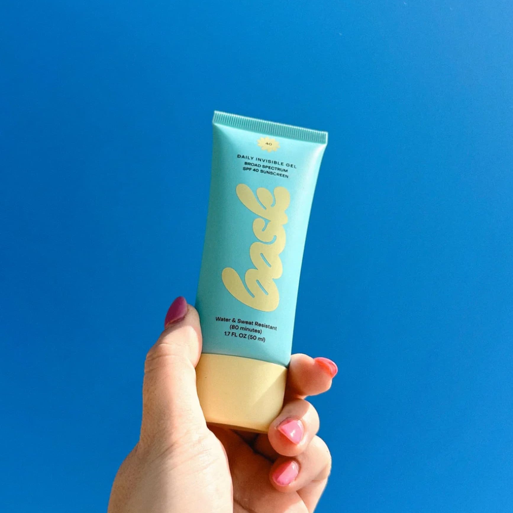 A Bask SPF 40 Face Sunscreen tube held in a hand against a blue sky.