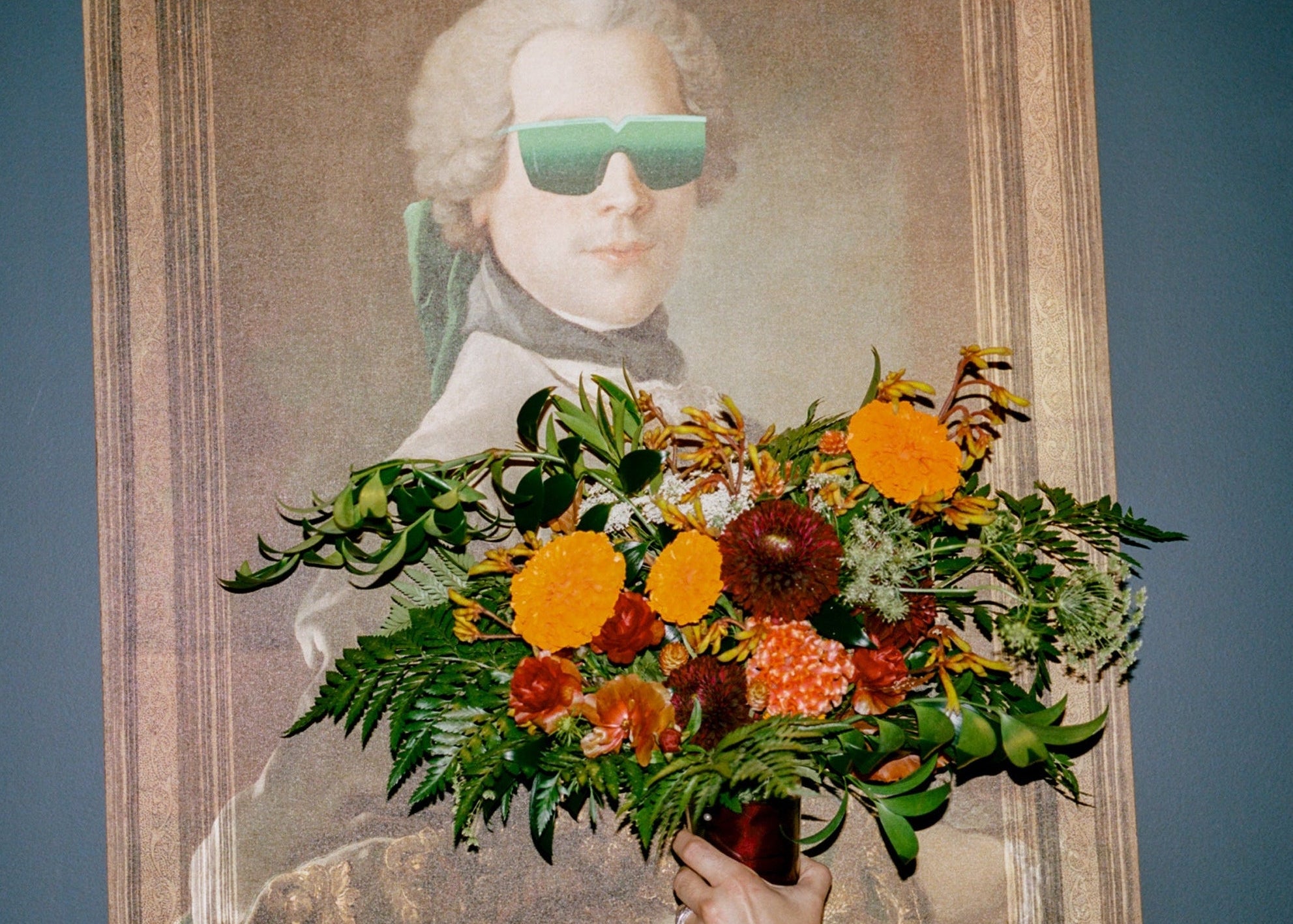 A hand holding a vibrant bouquet of orange and red flowers in front of a framed portrait painting of a man wearing green-tinted sunglasses, set against a dark blue wall.