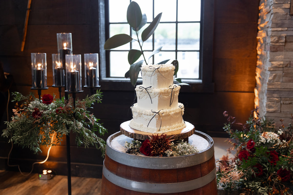 A three-tier white wedding cake with delicate black ribbon bows sits on a wooden barrel at a Diamond Mills wedding with warm candlelight, floral arrangements, and a window in the background.