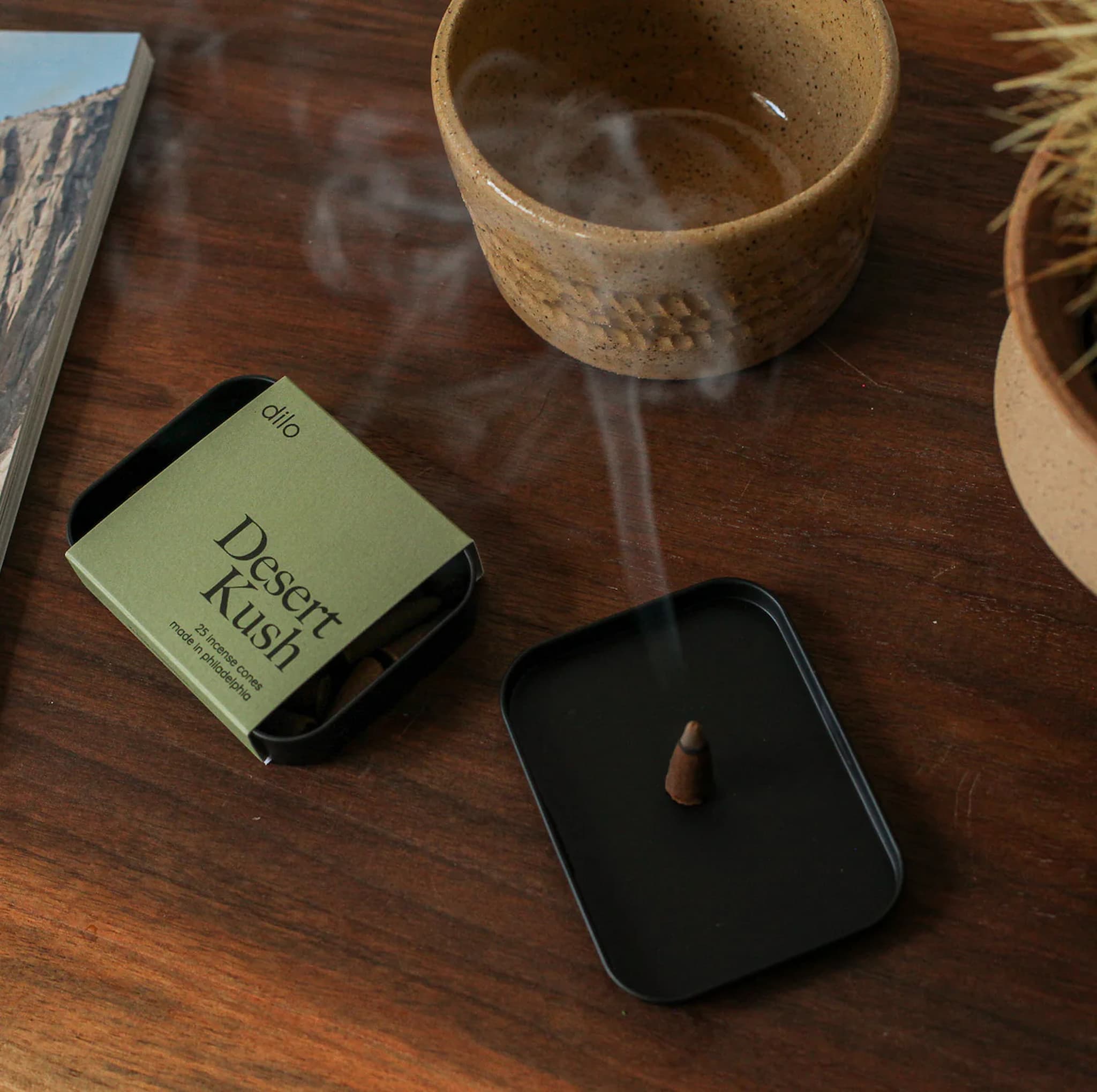 Dilo incense cone burning on a wooden table with a 'Desert Kush' packaging and a cup in the background.