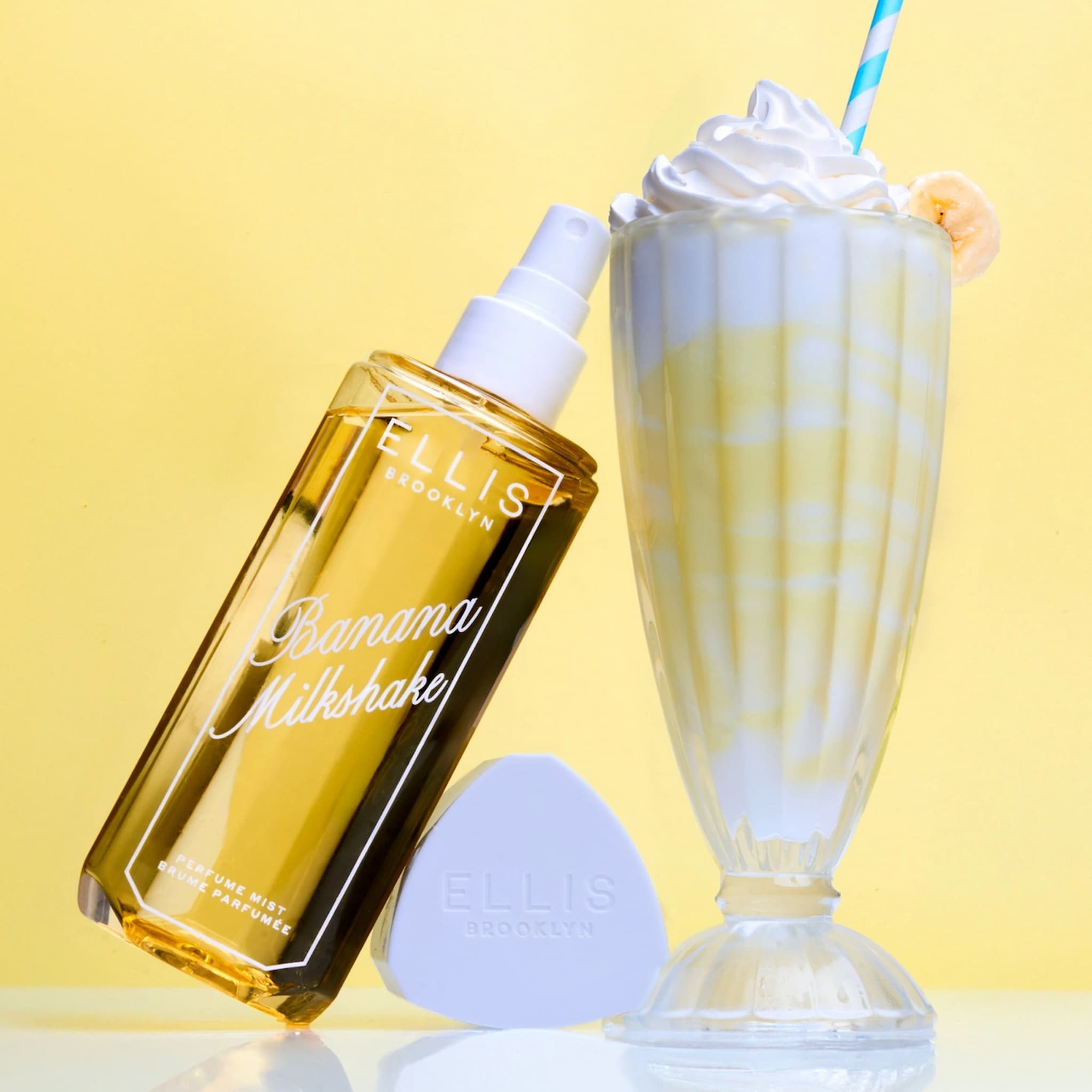 Ellis Brooklyn Banana Milkshake Perfume Mist tipped against a banana milkshake in a ridged clear glass with a straw.