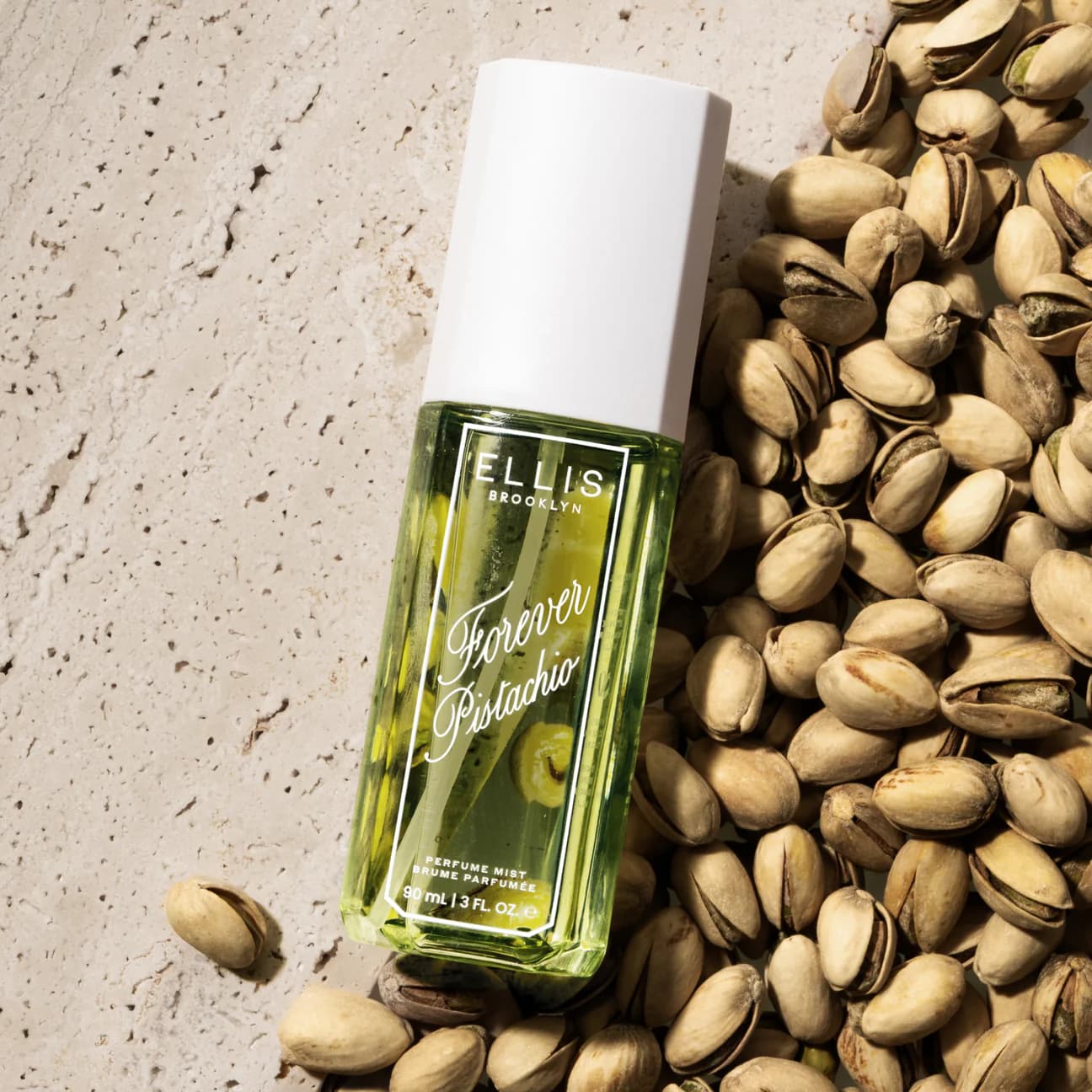 Bottle of Ellis Brooklyn Forever Pistachio perfume mist surrounded by pistachios on a textured surface.