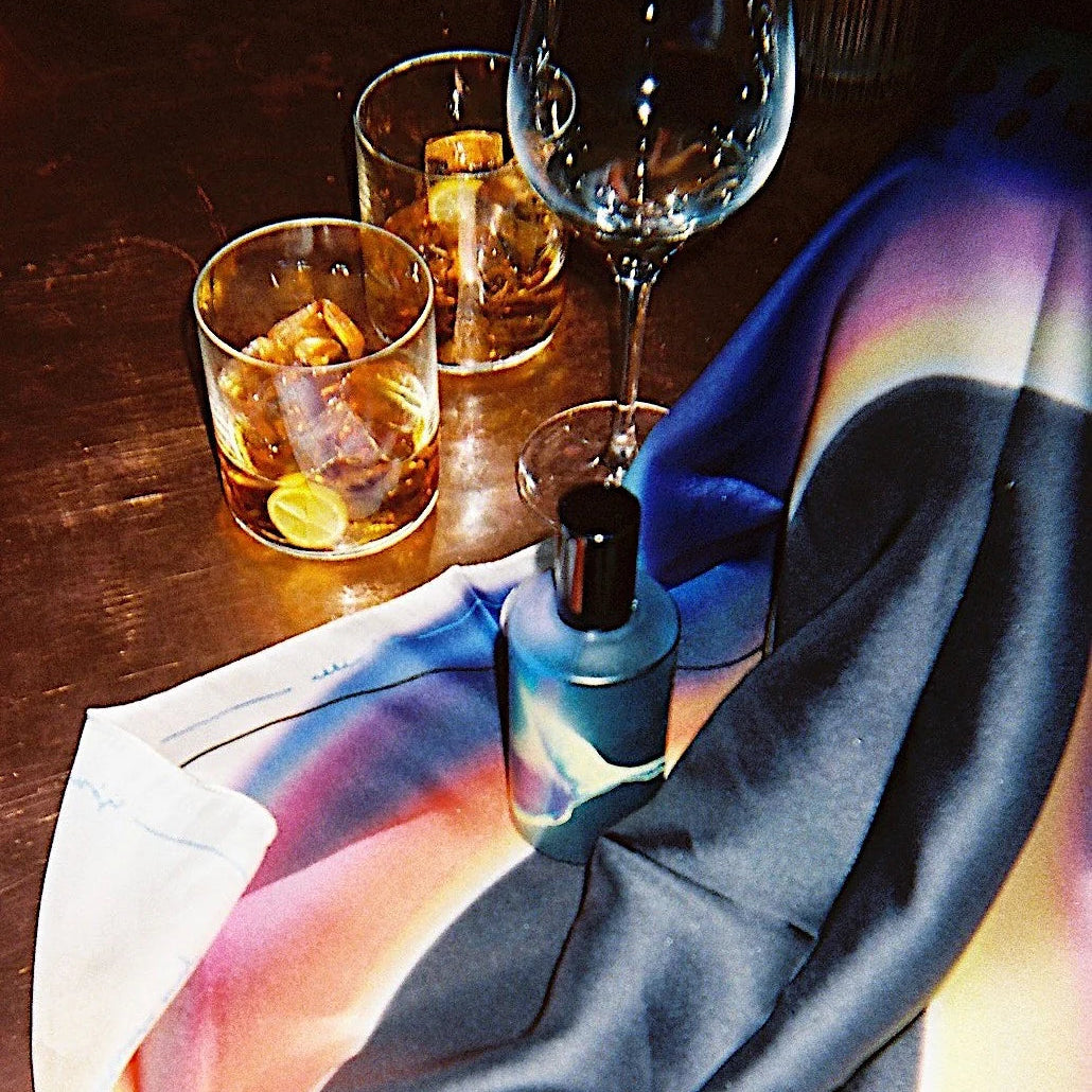 A Fischersund 4:45 bottle on a scarf with a colorful abstract sunset print in blue, purple, pink, and yellow, draped on a dark wooden table next to a wine glass, and two rocks glasses.