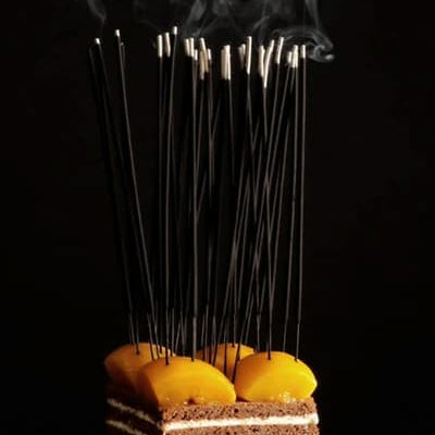 A layered cake topped with bright orange clementines, with many thin smoking Fischersund Jol incense sticks inserted into it, set against a dark background.