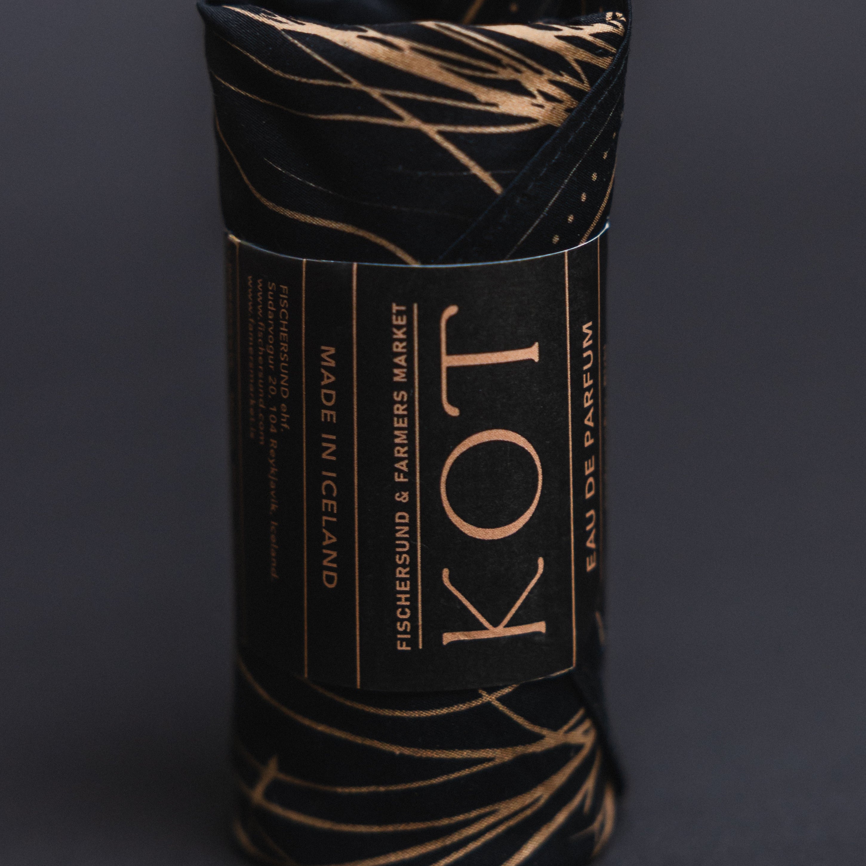 The Fischersund KOT fragrance rolled up in a black branded bandana with metallic gold abstract line patterns and a central paper band reading 'KOT' and 'EAU DE PARFUM', photographed on a dark gray background.