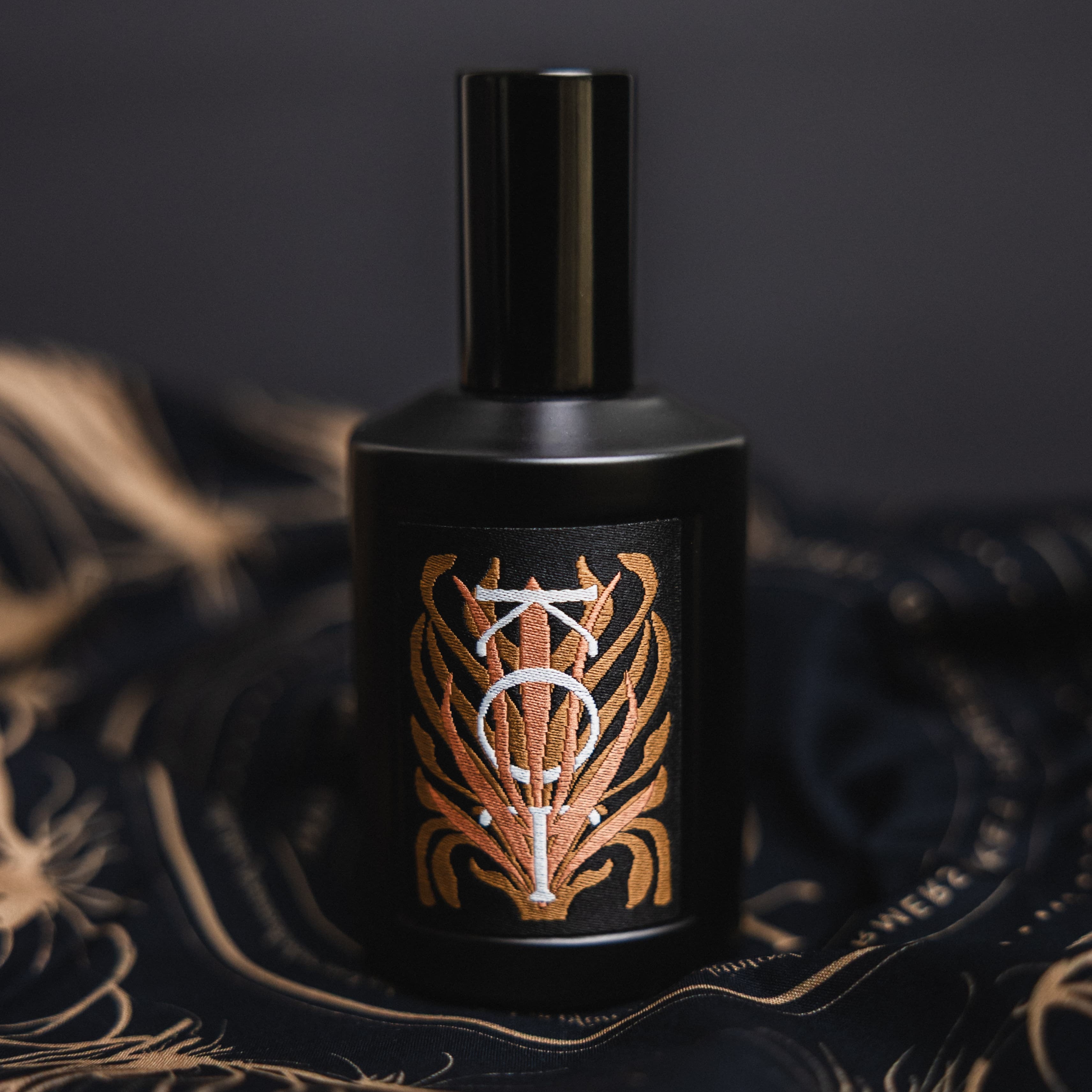 A black cylindrical bottle of Fischersund KOT perfume with a matching black cap, featuring a decorative label with a stylized white monogram over a gold pattern, set on a dark draped fabric with gold detailing.
