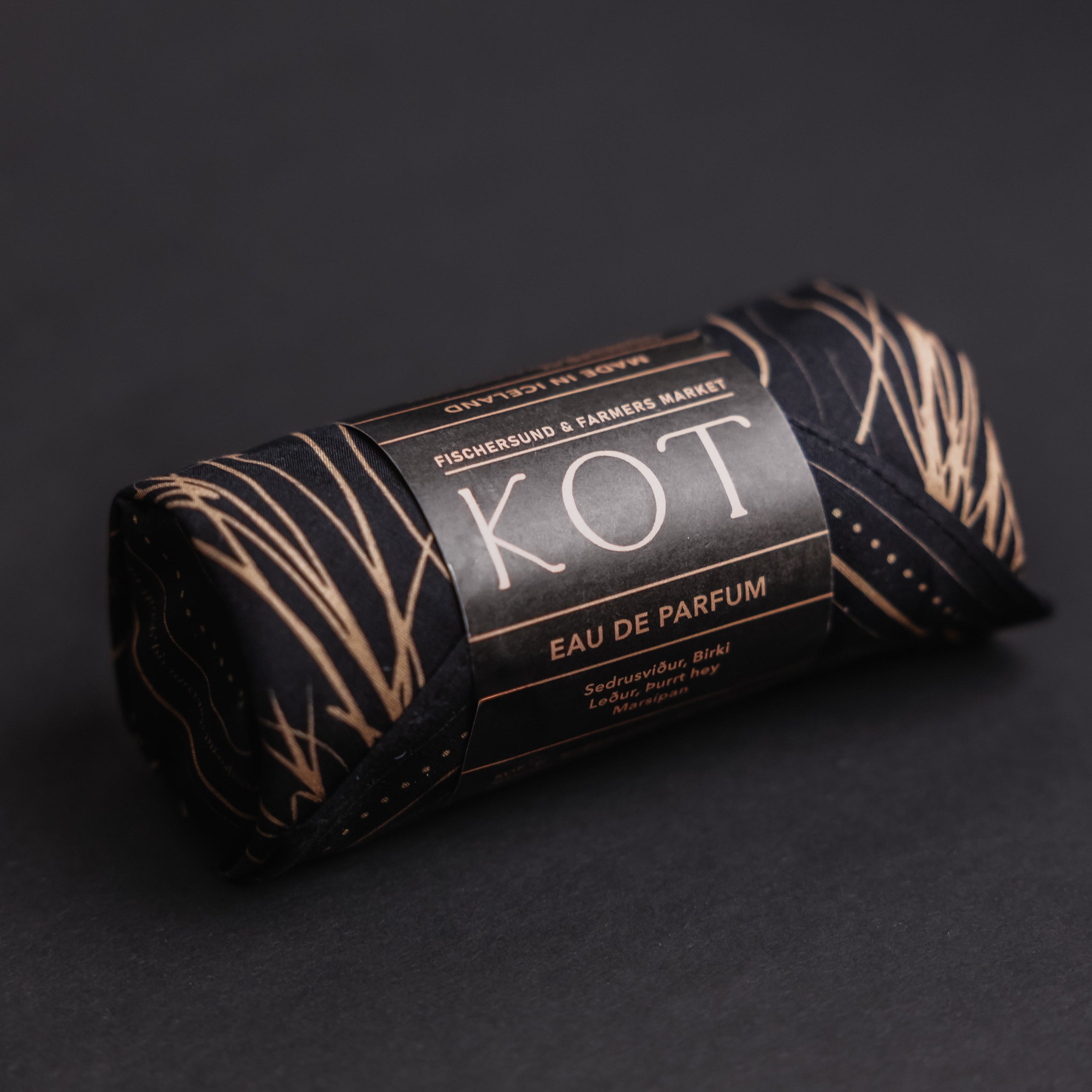A rolled-up black Fischersund KOT perfume bottle with a black paper label and black bandana with gold abstract line patterns, photographed on a dark gray background. The label reads 'KOT' and 'EAU DE PARFUM'.