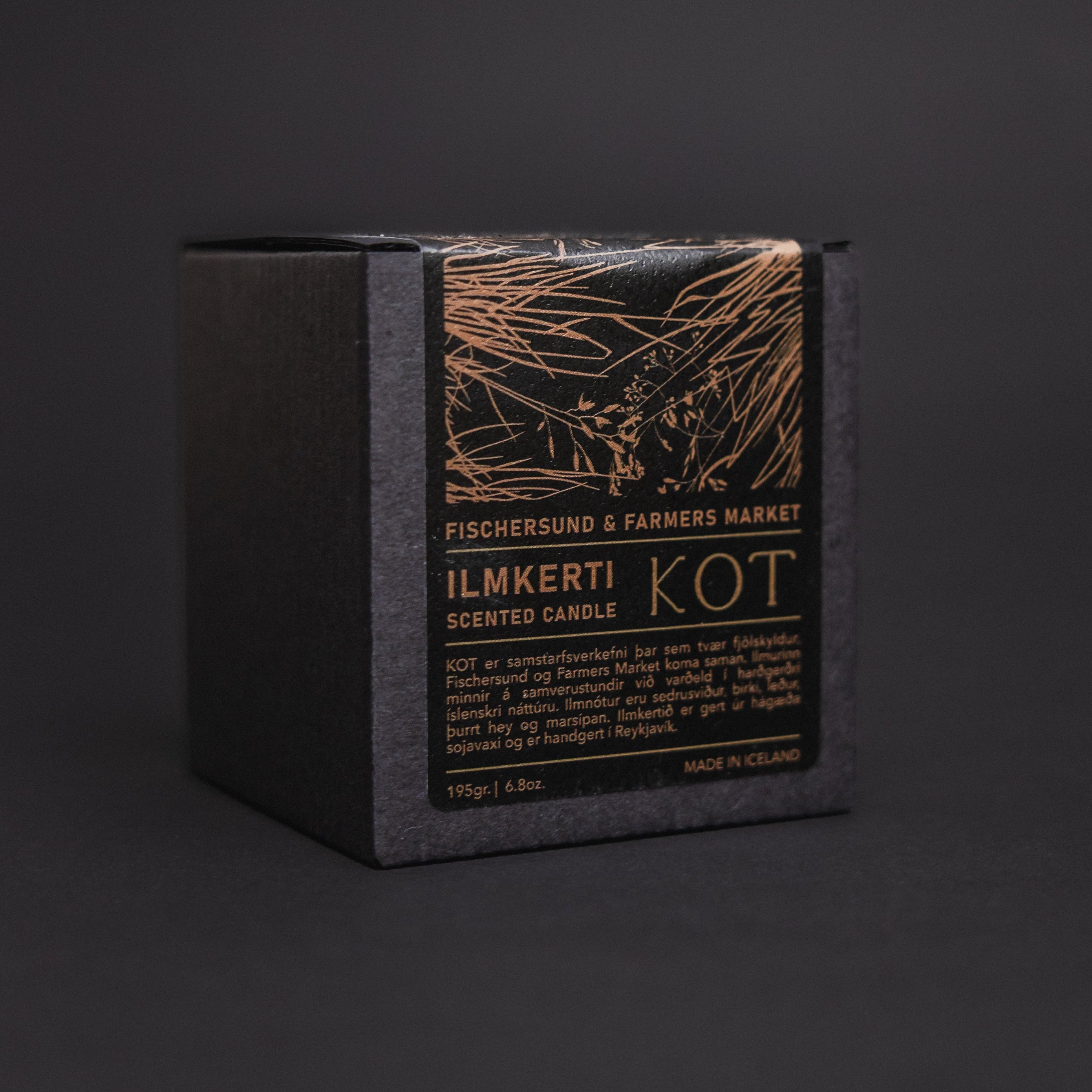 A black Fischersund KOT candle box with a black front label featuring gold text and an abstract gold line drawing, photographed on a dark gray background.