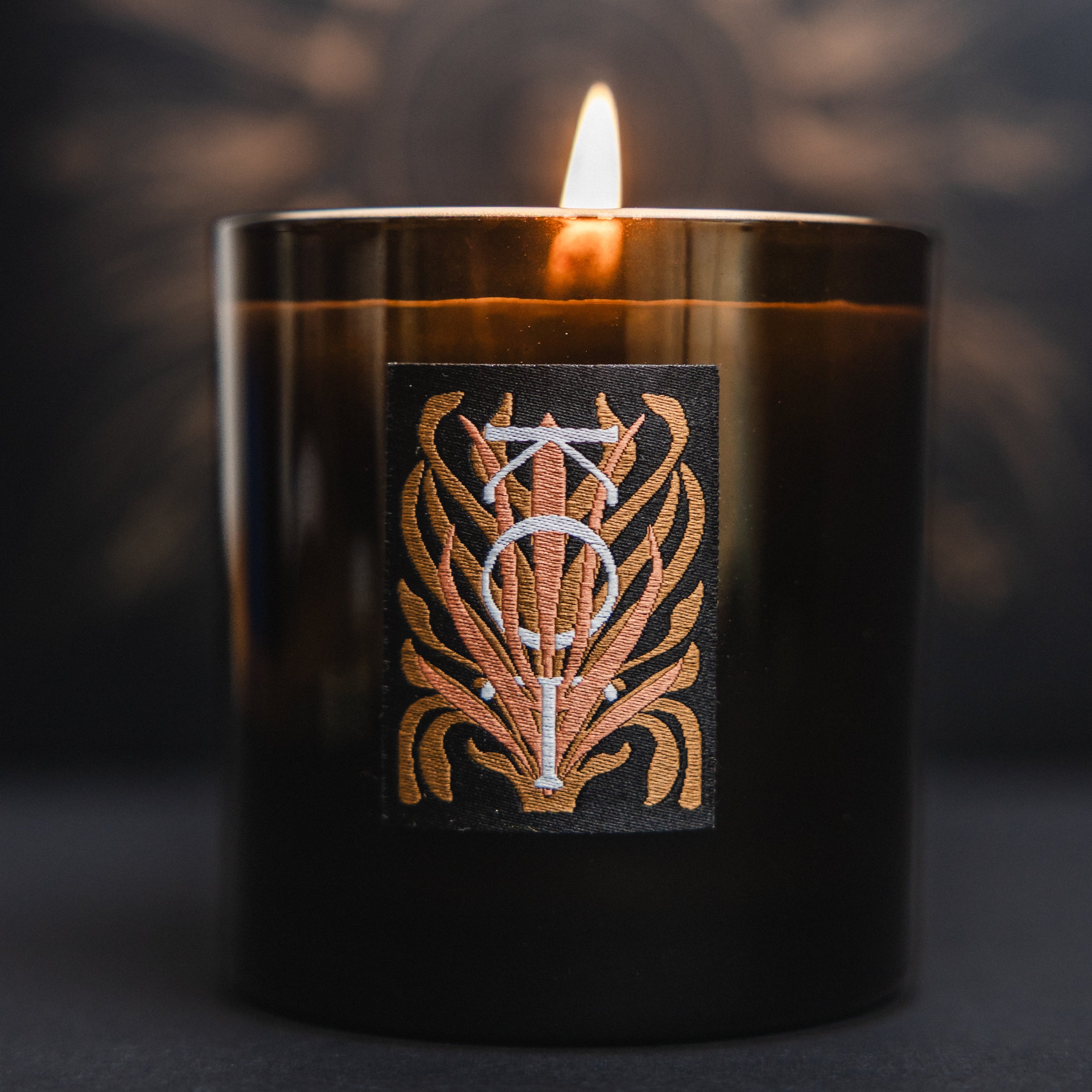 The Fischersund KOT black glass jar candle with a lit flame, featuring a decorative label with a stylized gold and white emblem, set against a dark background with a soft sunburst glow.