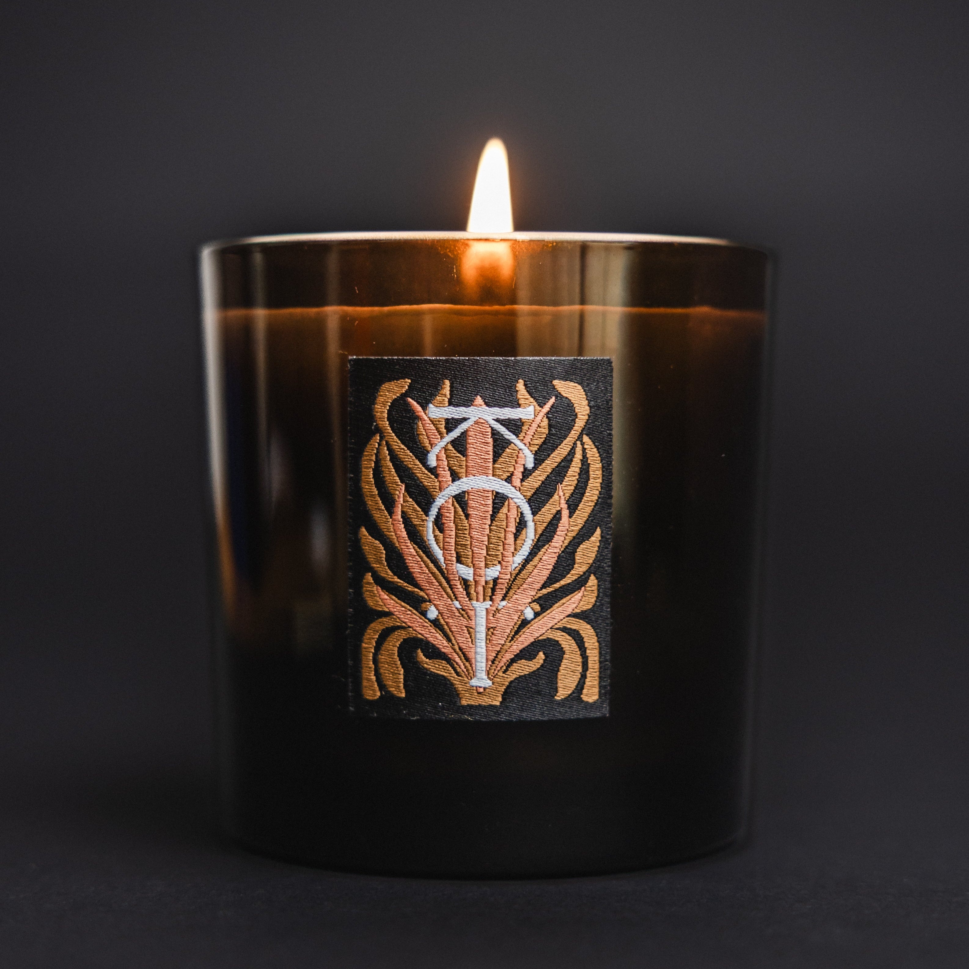 A lit Fischersund KOT black glass candle jar with a warm amber cast, featuring a rectangular black label with a metallic gold and white ornate emblem, set against a dark gray background.