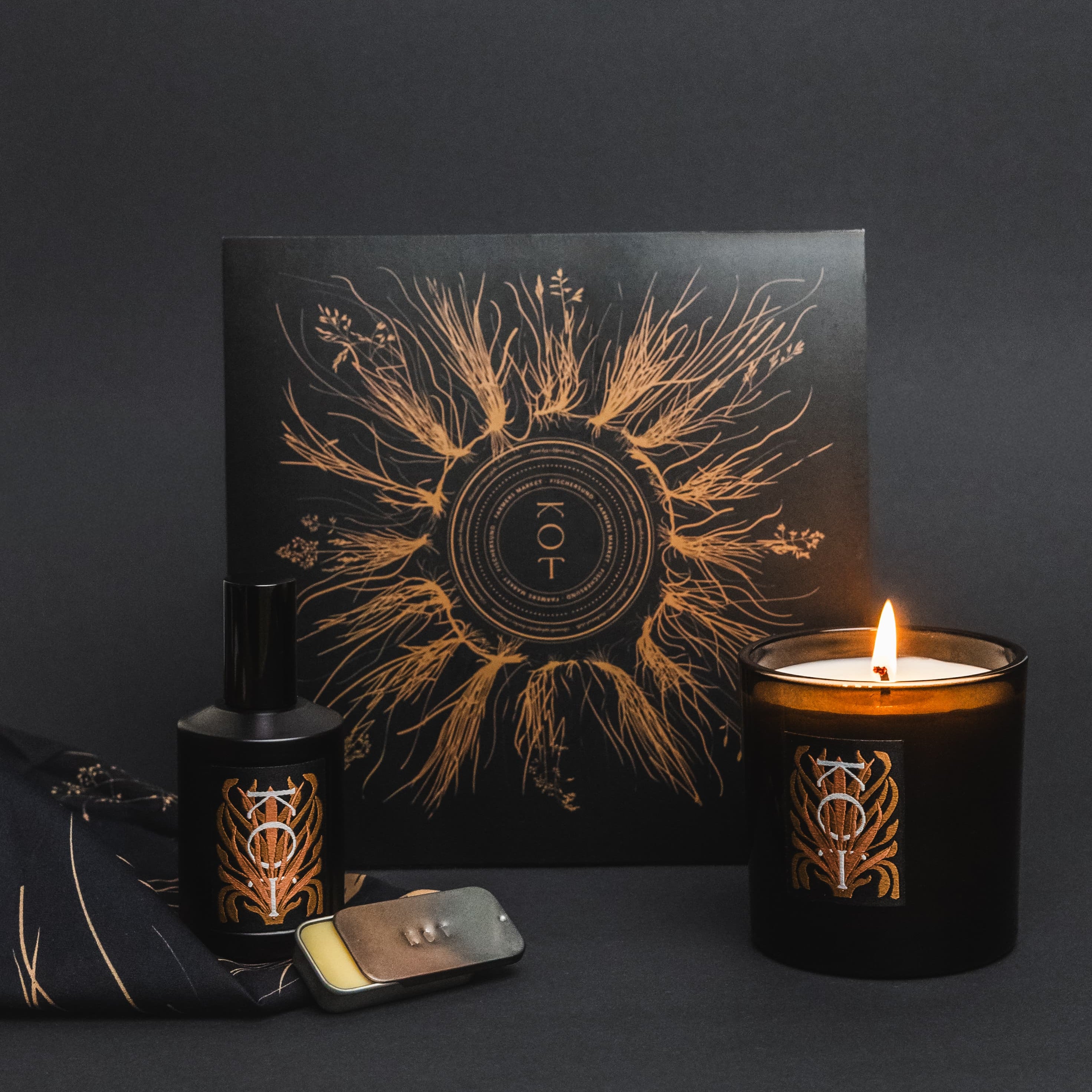 A Fischersund x Farmers Market KOT colletion set featuring a black jar candle with a lit flame, a black bottle of KOT perfume, a folded black bandana, and a square box with a gold sunburst and circular symbol, arranged on a dark surface against a dark background.