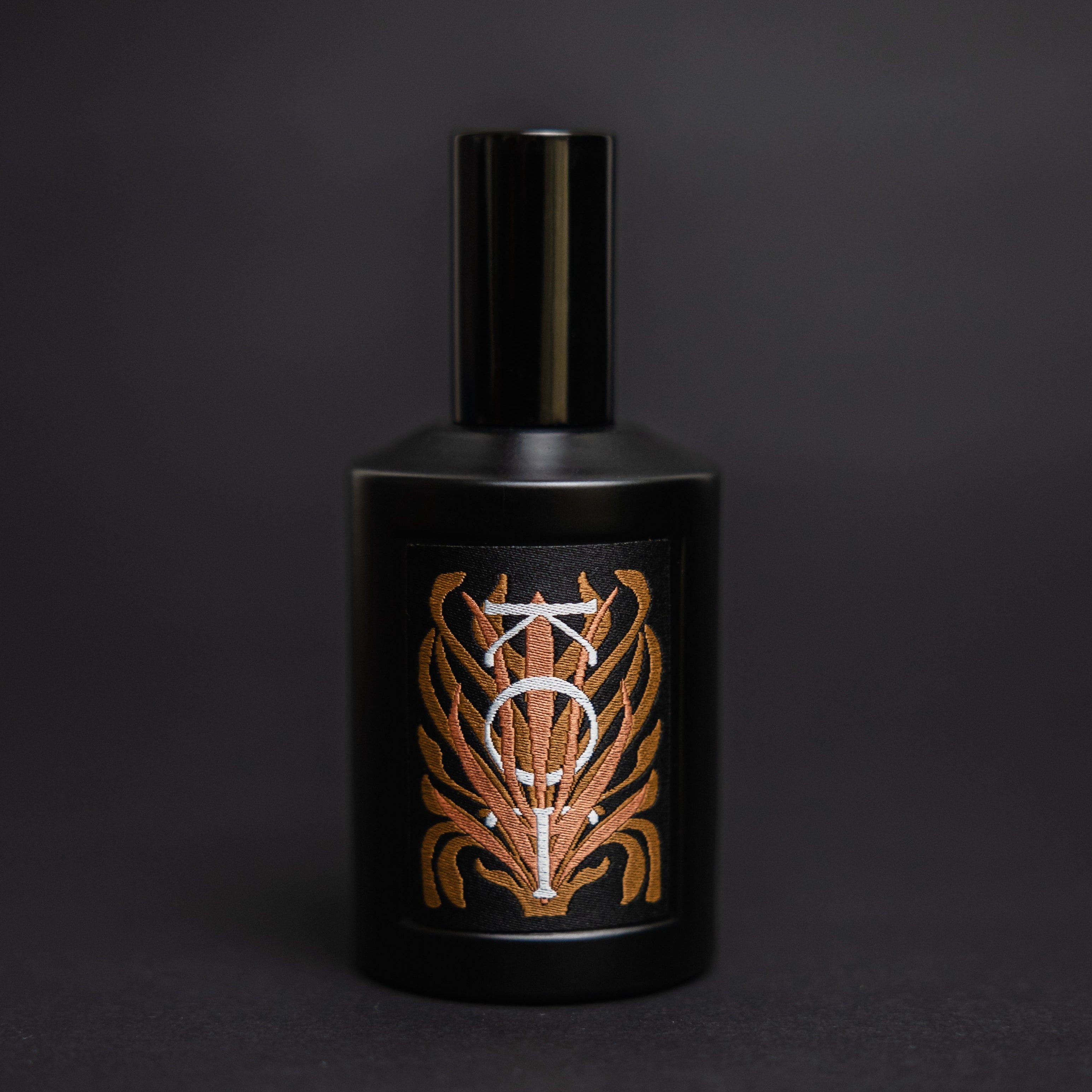 A matte black cylindrical bottle of Fischersund KOT fragrance with a matching black cap, featuring a front label with a stylized metallic gold and white emblem, set against a dark gray background.