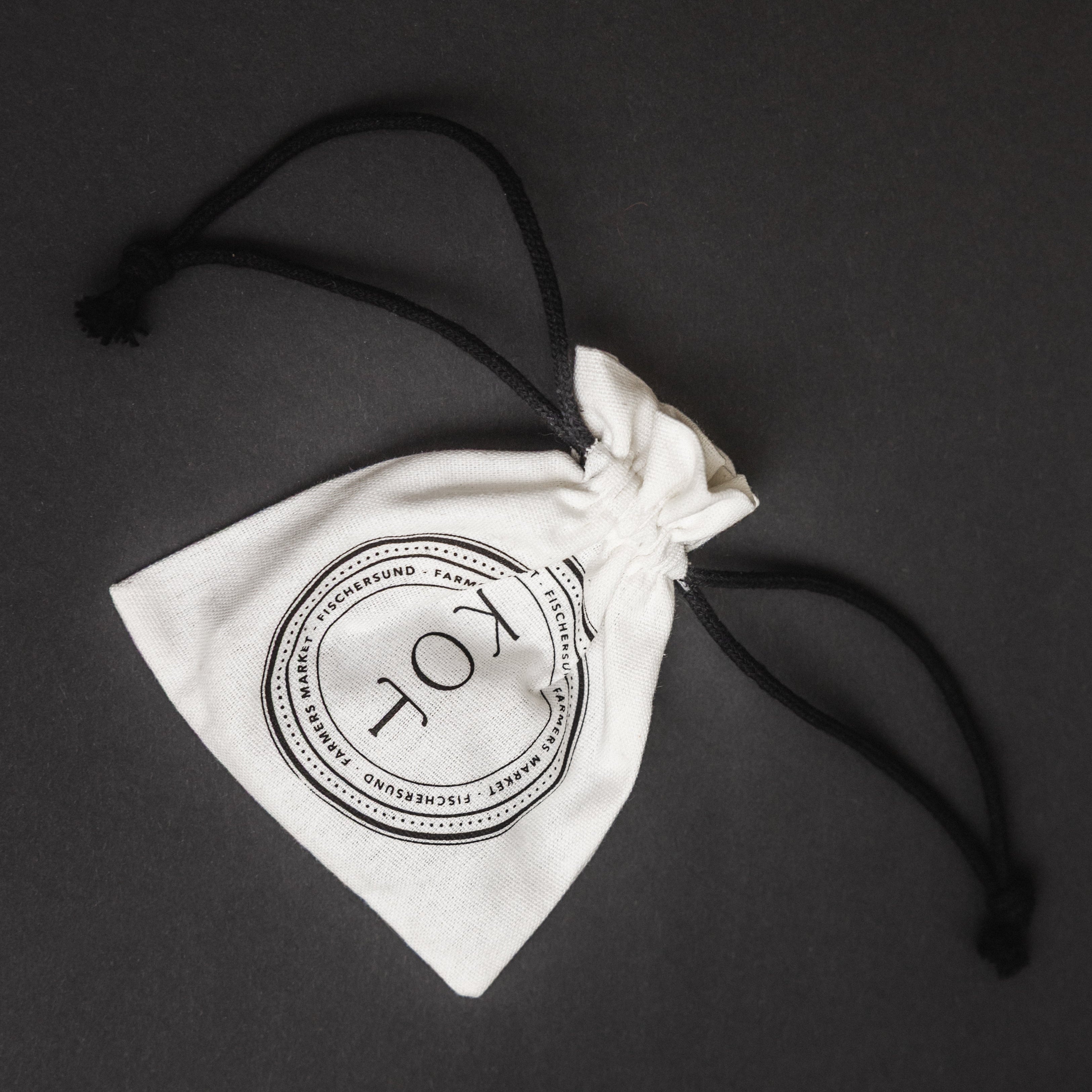 A white cotton drawstring pouch with a black printed circular logo reading 'KOT' and black drawstring cords, photographed on a dark gray background.