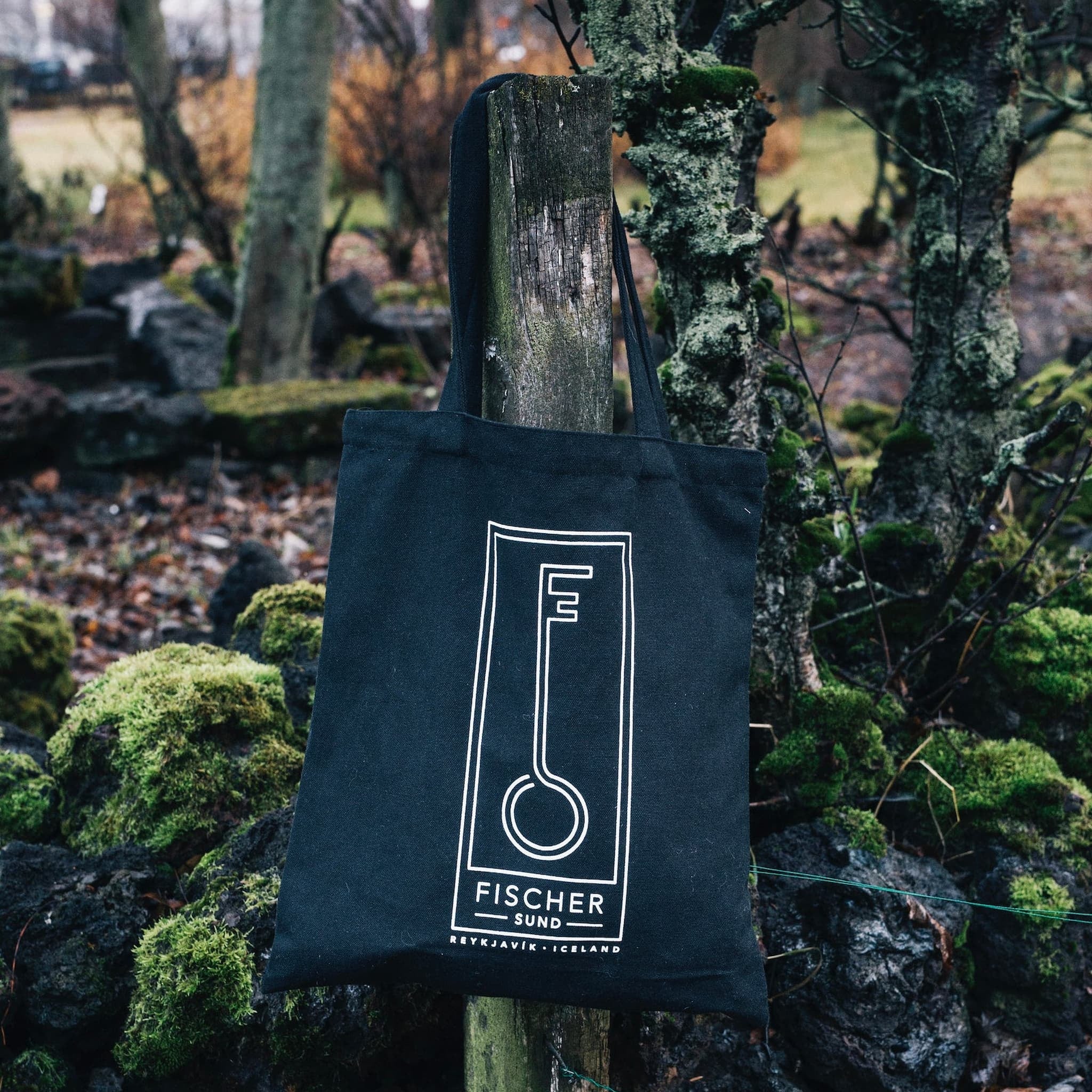Black Fischersund tote bag with a logo and text on a wooden post in a natural setting