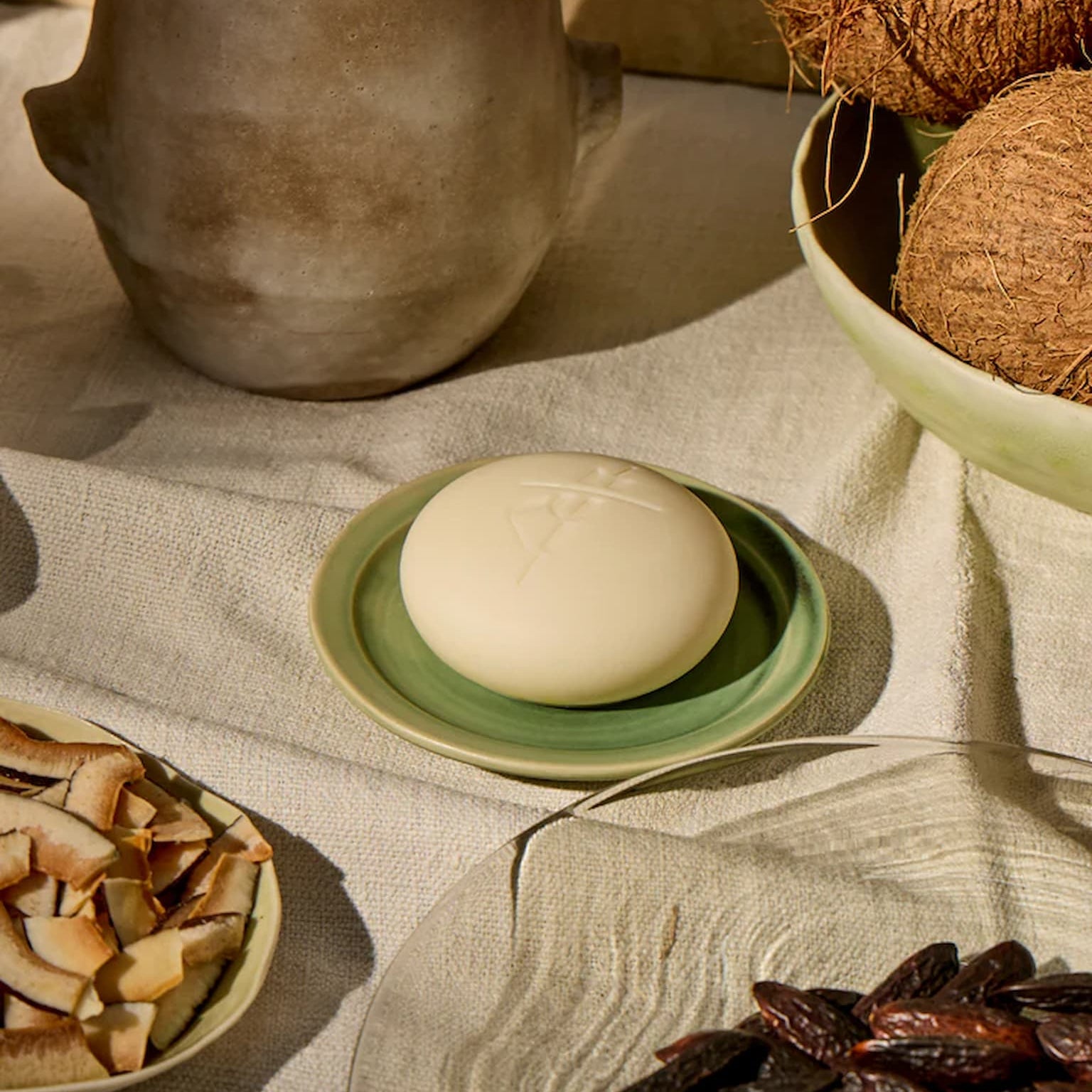 Kate McLeod Body Stone in Limited Edition Tonka Bean & Coconut scent, on a green saucer on a beige tablecloth with coconuts in the background.