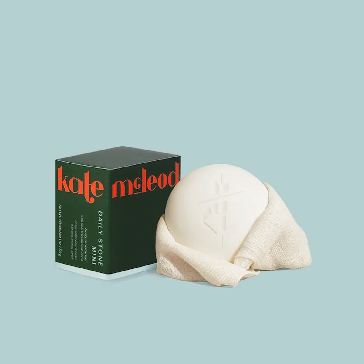 A white bar of the mini Kate McLeod Daily Stone sits wrapped in a cream-colored fabric, next to the green product packaging.
