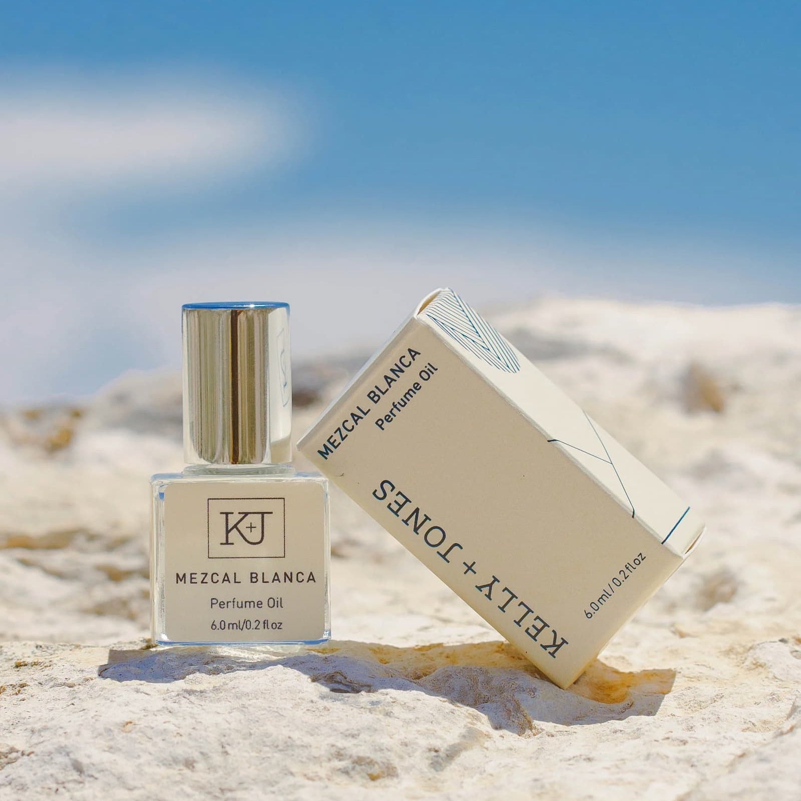 Kelly + Jones Mezcal Blanca perfume oil bottle and packaging on a sandy beach surface with a blue sky.