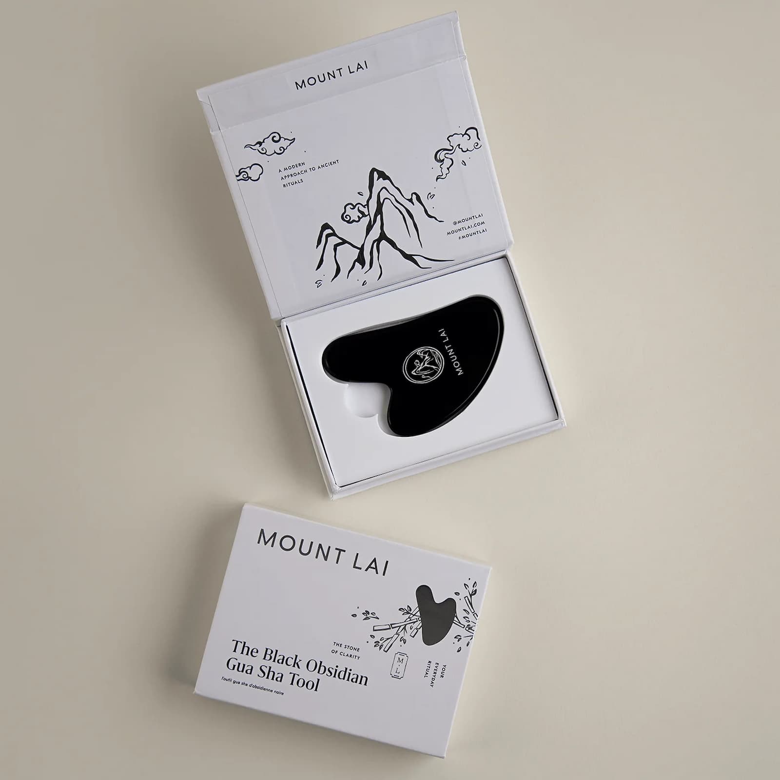 A Mount Lai Obsidian Gua Sha tool inside of its white rectangular packaging, against a grey-beige background