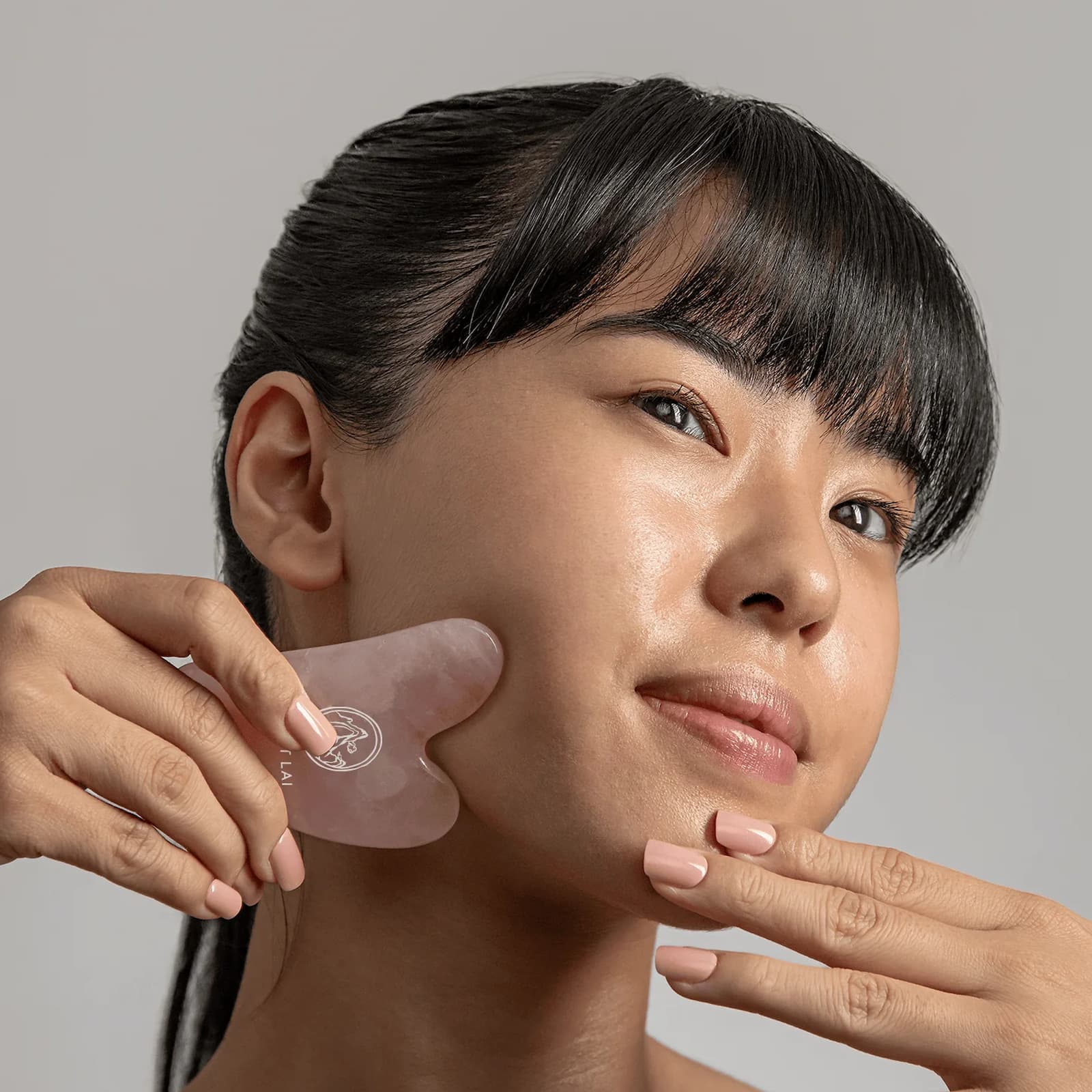 A woman with dark hair and bangs uses a translucent pink Mount Lai Rose Quartz Gua Sha tool on her cheek, with her other hand touching her chin, against a solid gray background.