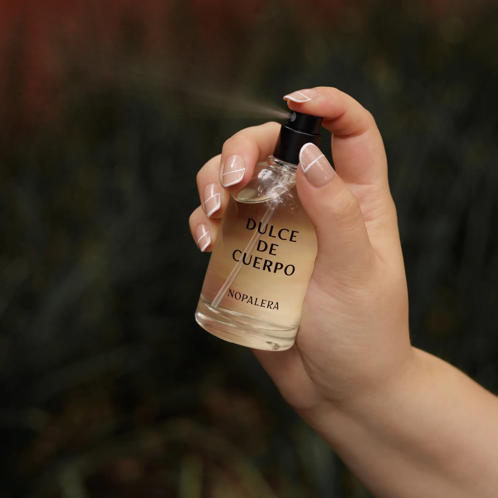 A hand is spraying a glass bottle of Nopalera Dulce de Cuerpo perfume against a green background.