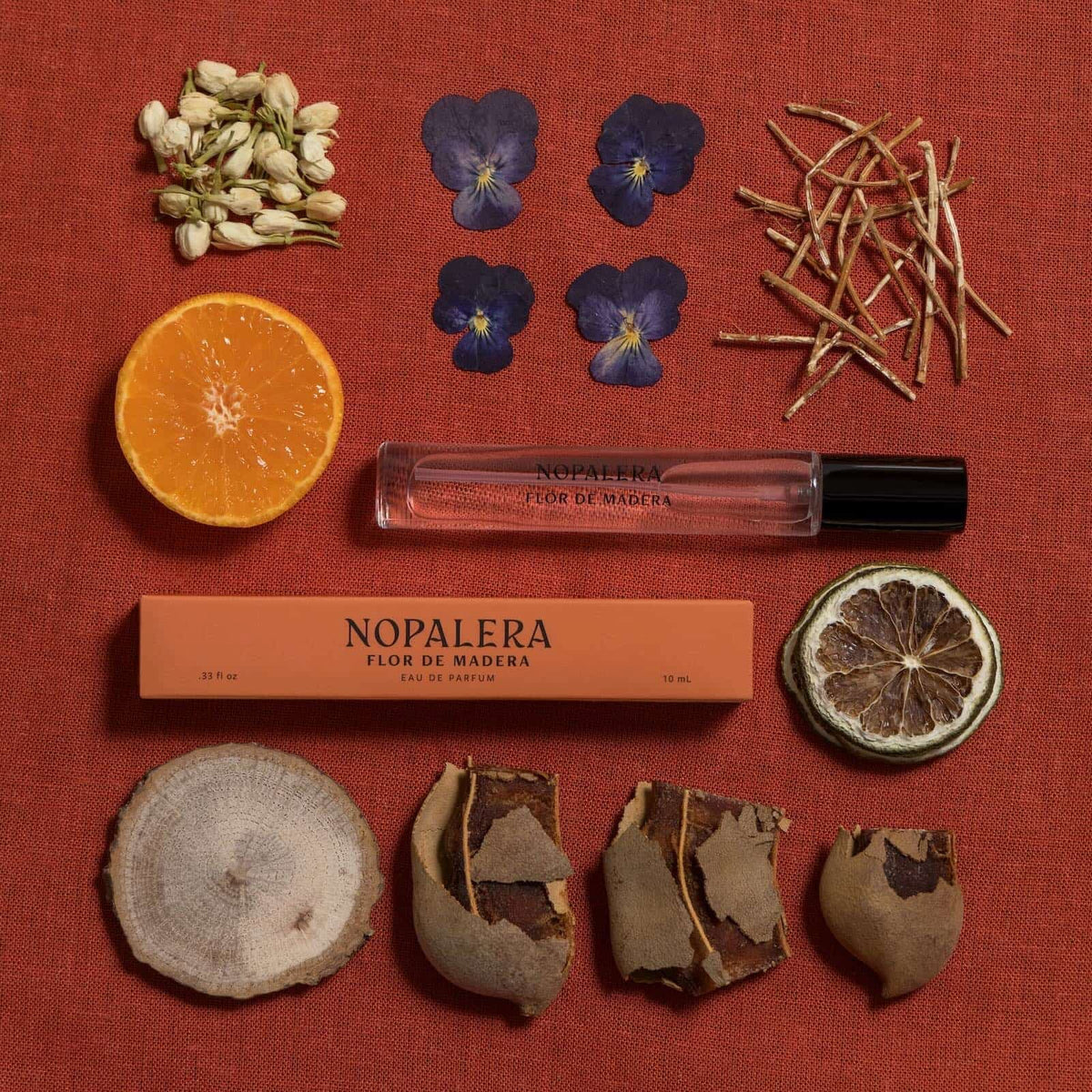 A travel-sized glass bottle of Nopalera "Flor de Madera" perfume and its packaging laid flat against a dark orange background. It is surrounded by fragrance ingredients, including citrus, violet, jasmine, and cedarwood.