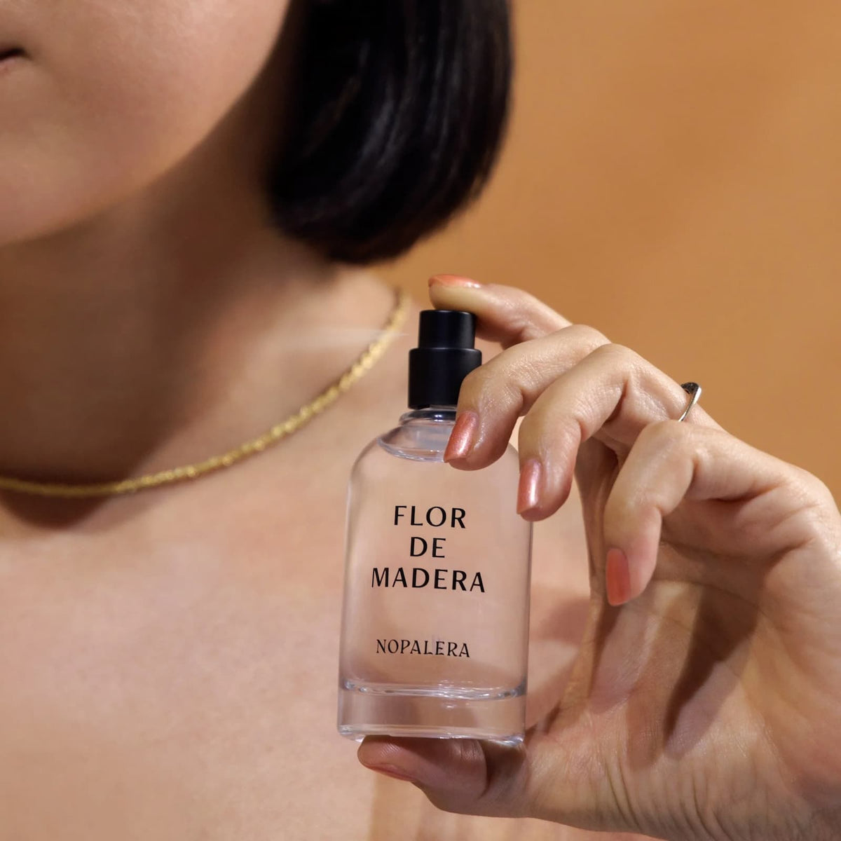 A person holding a glass bottle of Nopalera "Flor de Madera" perfume, spraying it against their neck.