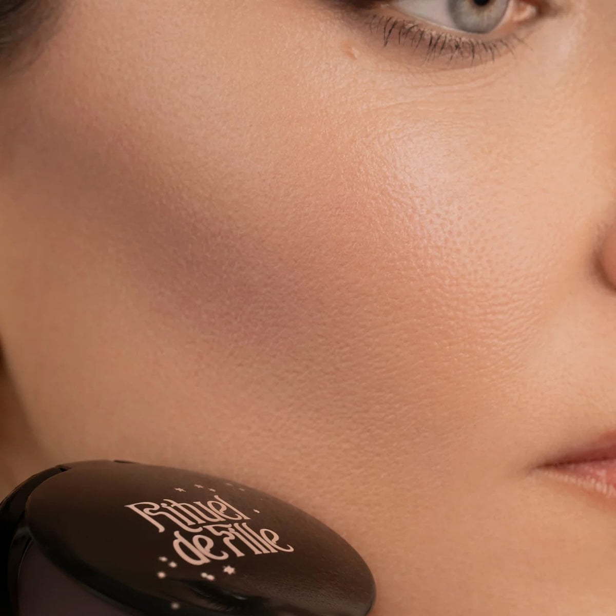 A close-up of a model's face, holding a makeup compact to her cheek. They are wearing Rituel de Fille Inner Glow Creme Pigment in "Intuition".