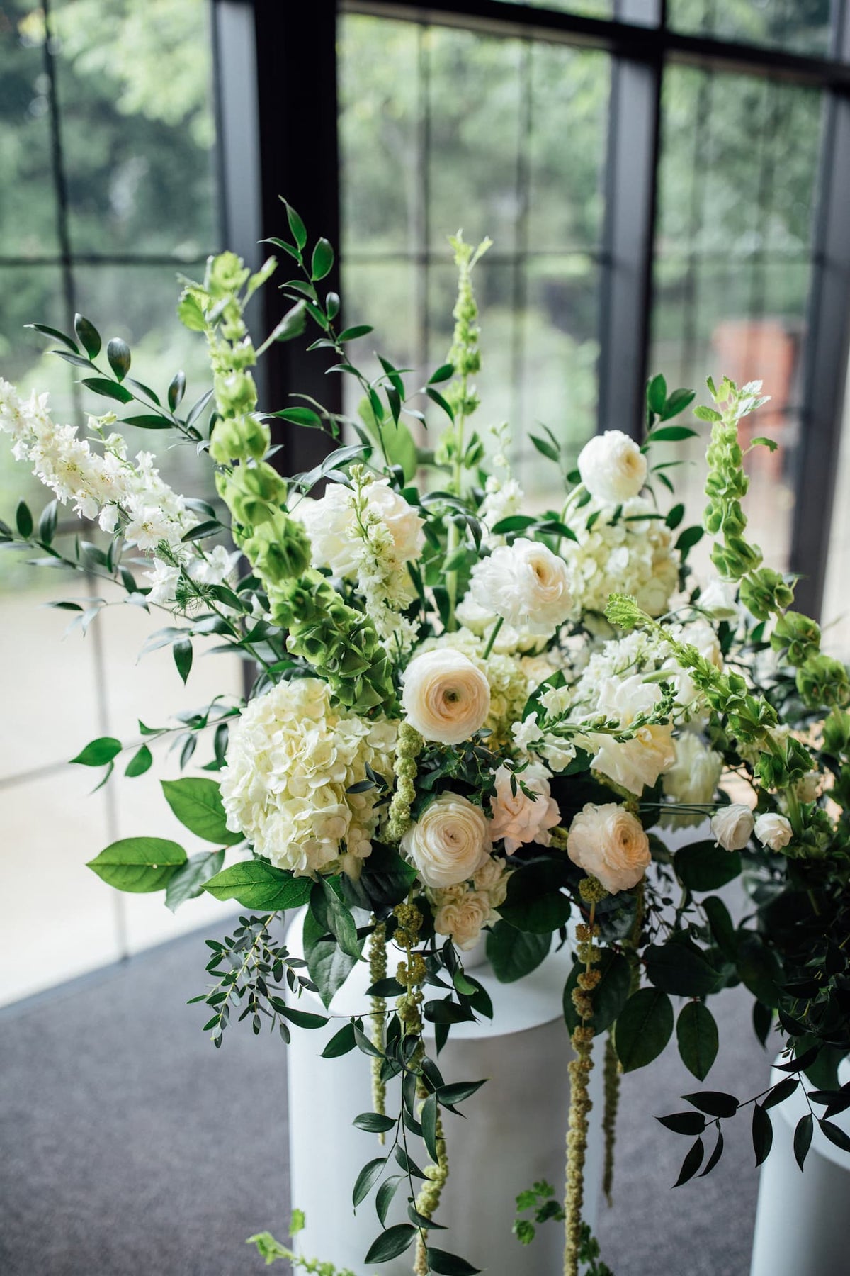 A large floral arrangement at a Roundhouse wedding featuring cream and white flowers with green foliage, displayed on a white pedestal in front of a window.