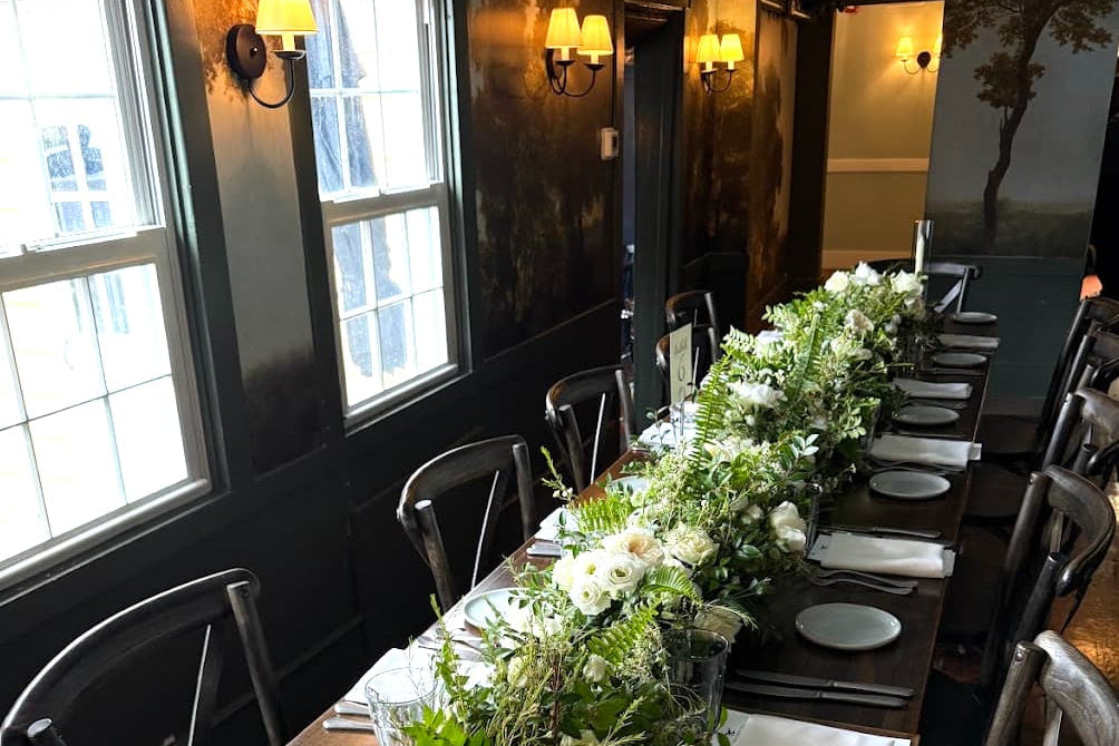 A long dining table at a Bird & Bottle Inn wedding in Garrison, NY, featuring white floral centerpieces with green foliage, white plates, folded white napkins, and clear glass candle holders.