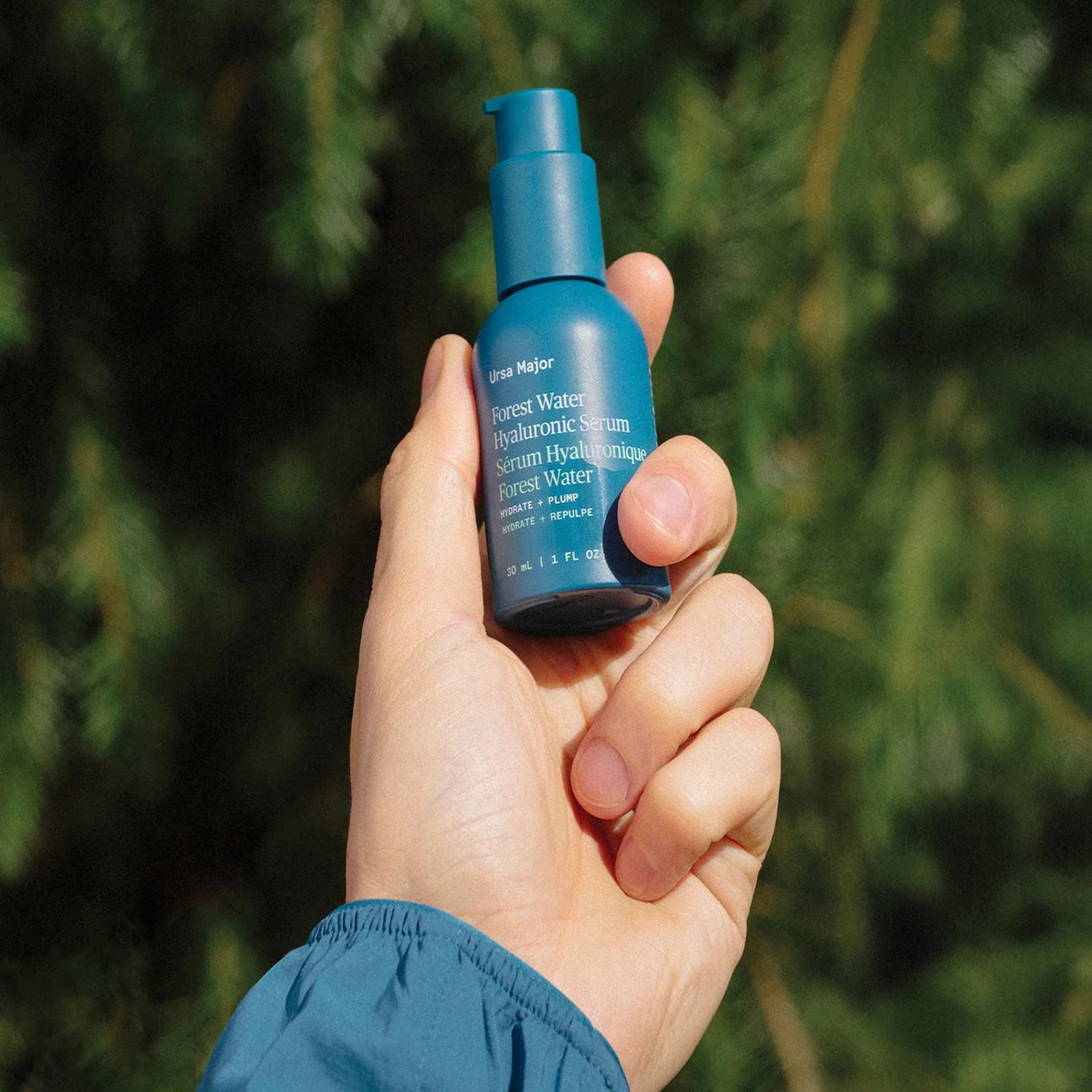A hand is holding a blue bottle of Ursa Major Forest Water Hyaluronic Serum against a background of green pine leaves.