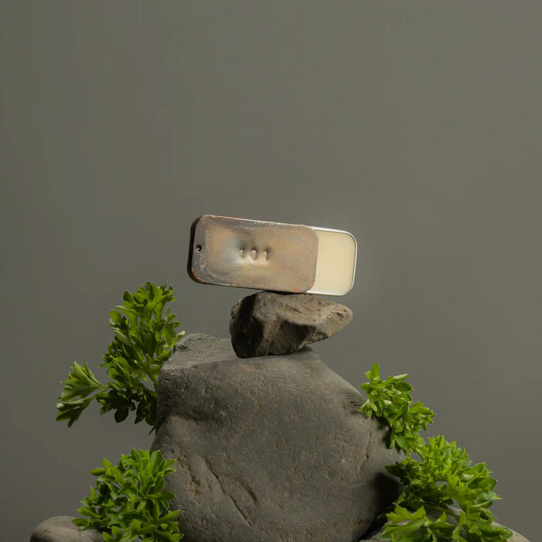 Balanced rock sculpture with a tin of Fischersund solid perfume on a gray background