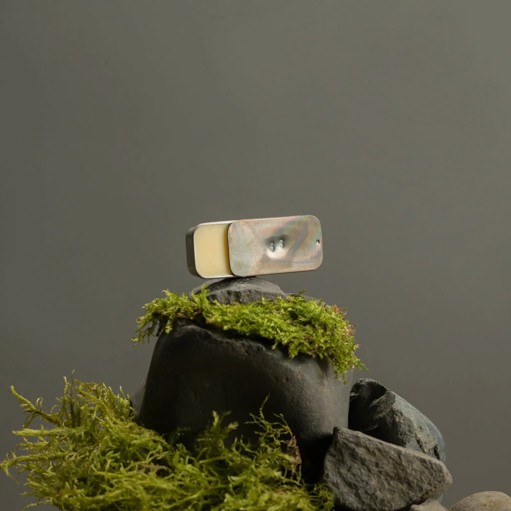 Metallic tin holding Fischersund solid perfume balanced on a rock with green moss against a gray background