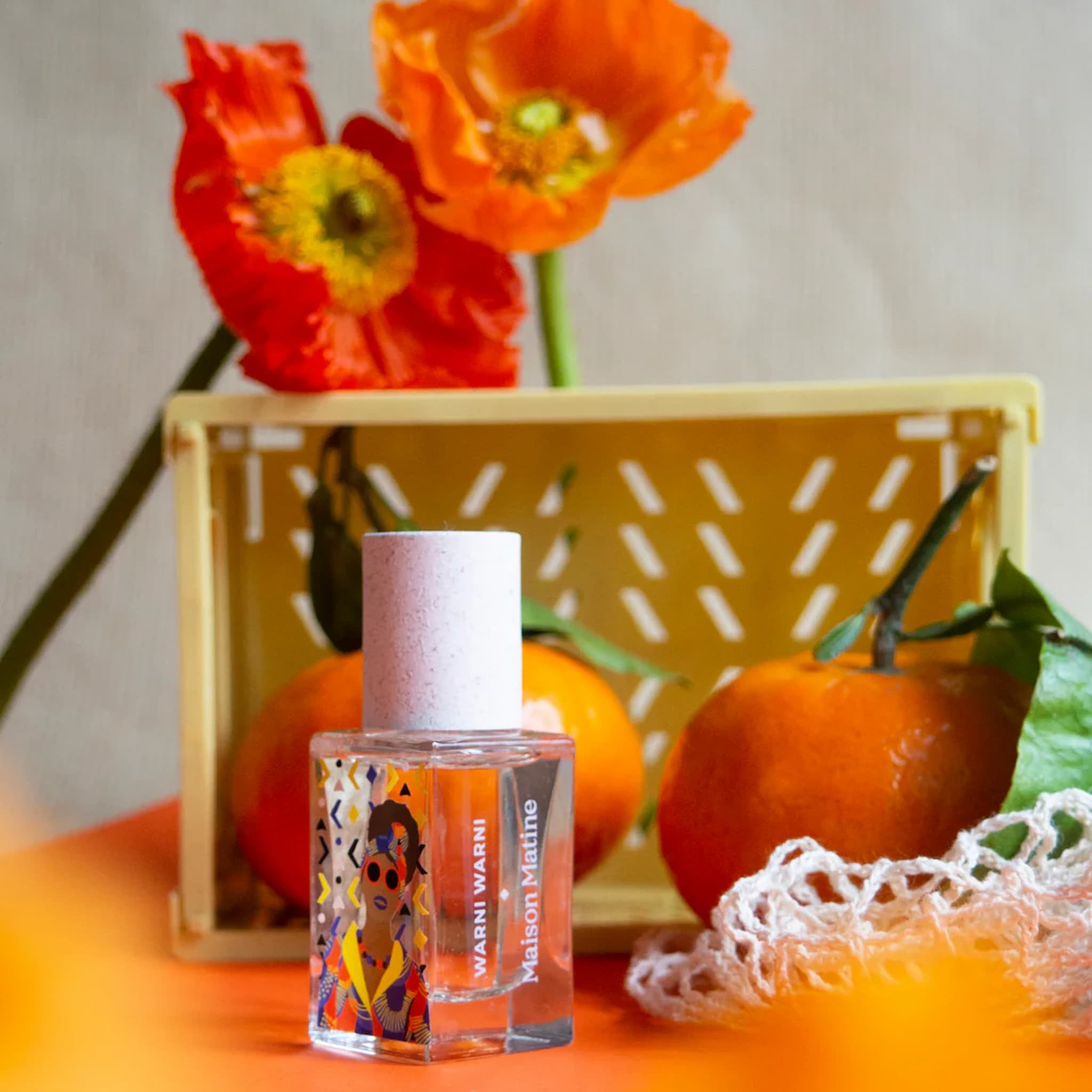 Small bottle of perfume with colorful label on an orange surface, surrounded by oranges and flowers.