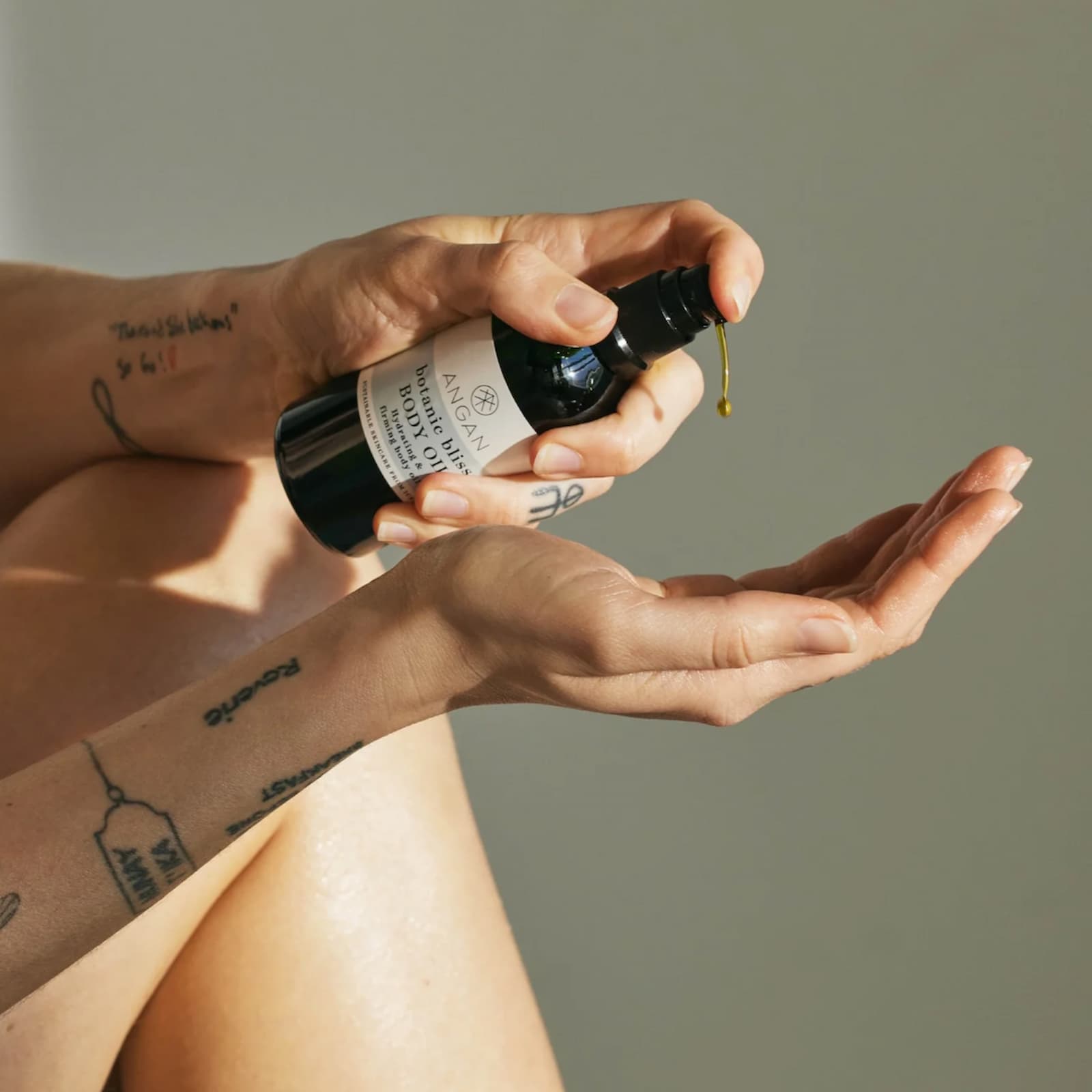 One hand is dispensing Angan Westfjords Botanic Bliss Body Oil into the other, against a grey-green background.