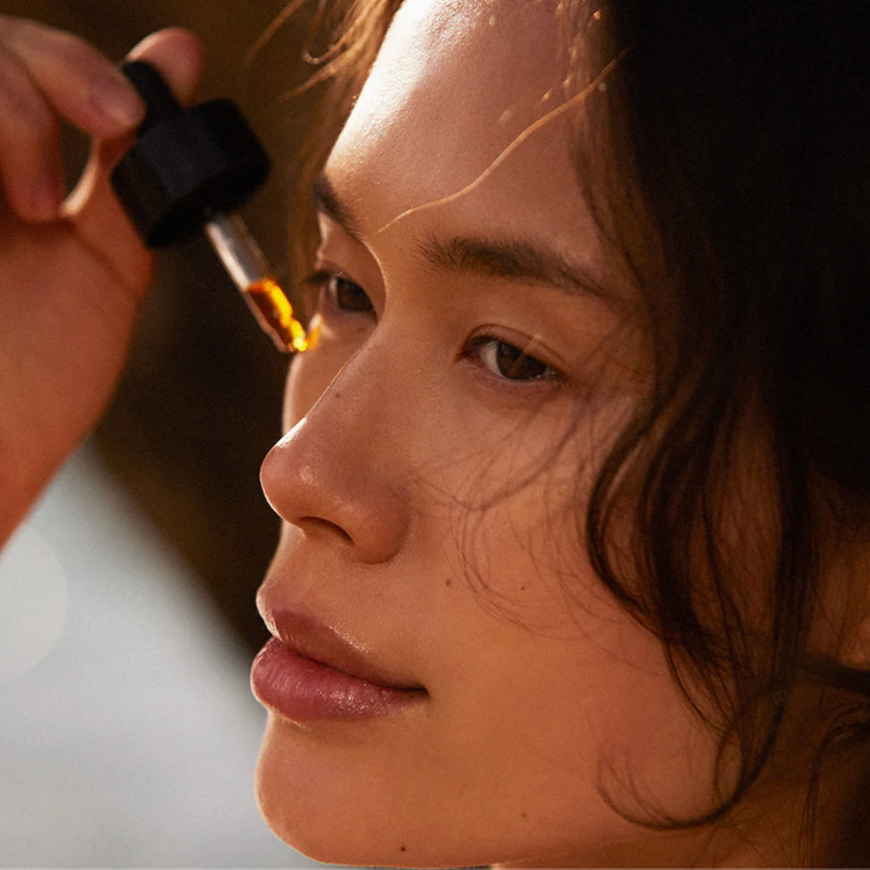Close-up of a person applying a dropper of Angan oil serum to the cheek.
