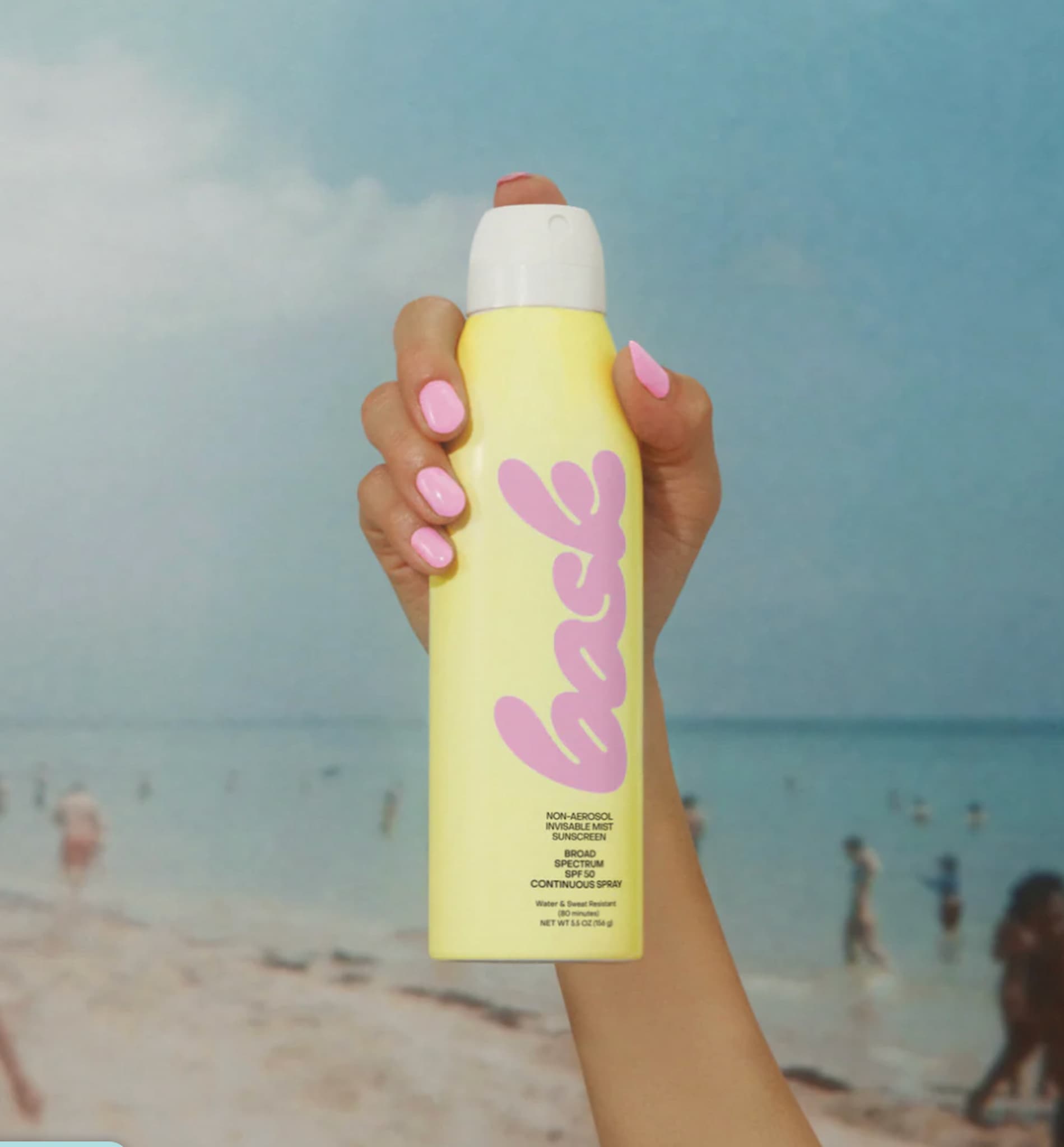 Bask SPF 50 Non-Aerosol Sunscreen Spray bottle in a hand, spraying into the air on a beach background.