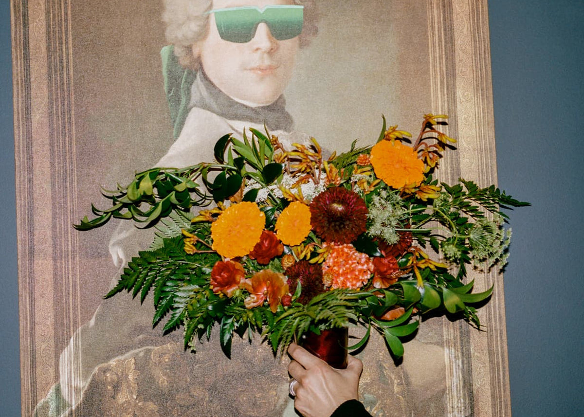 A Bird & Bottle wedding bridal bouquet. A hand holds a vibrant bouquet of orange, yellow, and red flowers in front of a framed portrait painting of a man wearing green-tinted sunglasses, set against a dark blue wall.