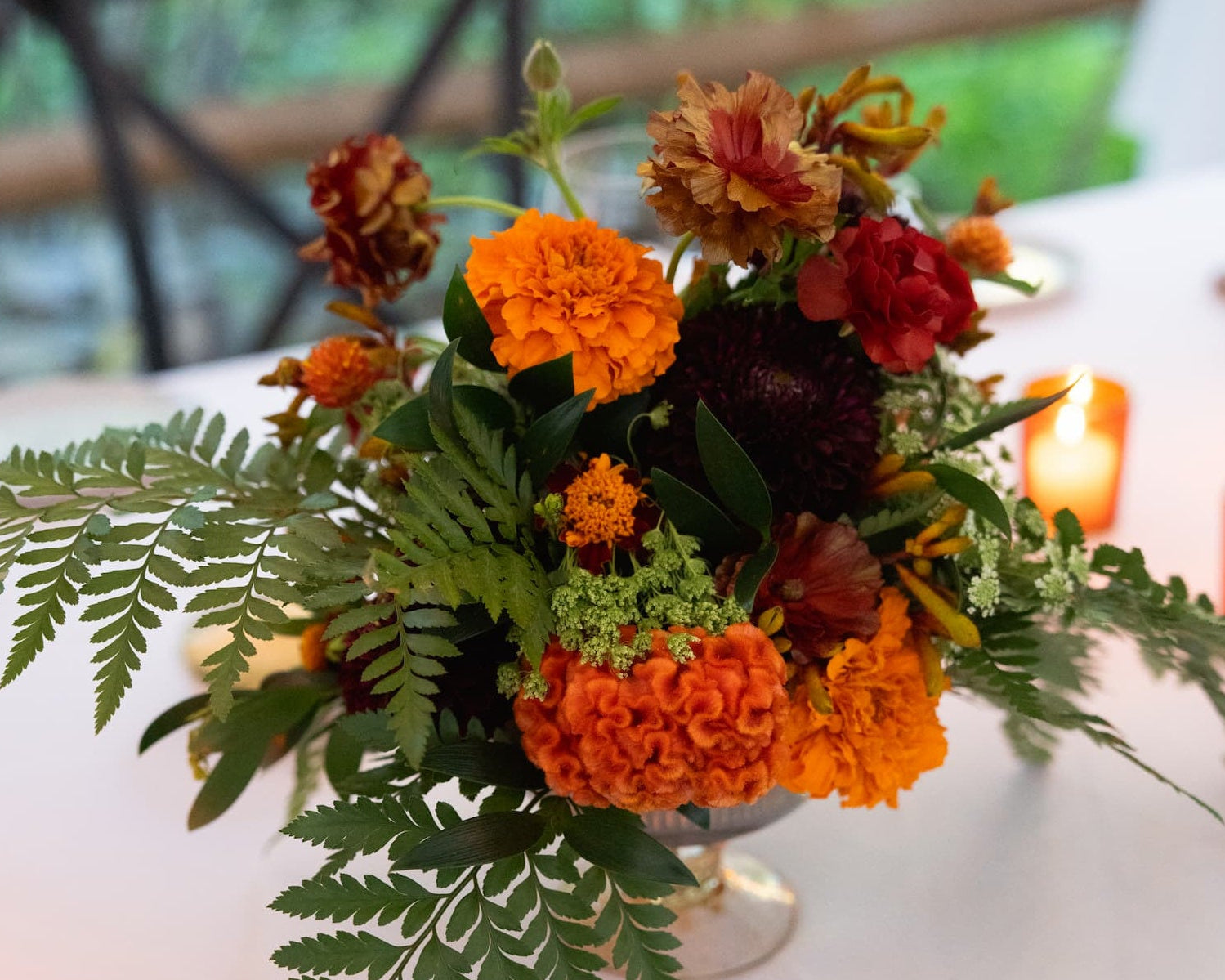 An autumn floral centerpiece at the Bird & Bottle Inn in Garrison, NY, in an amber bowl, featuring vibrant orange marigolds, deep red ranunculus, and green ferns, set on a white tablecloth with warm candlelight in the background.
