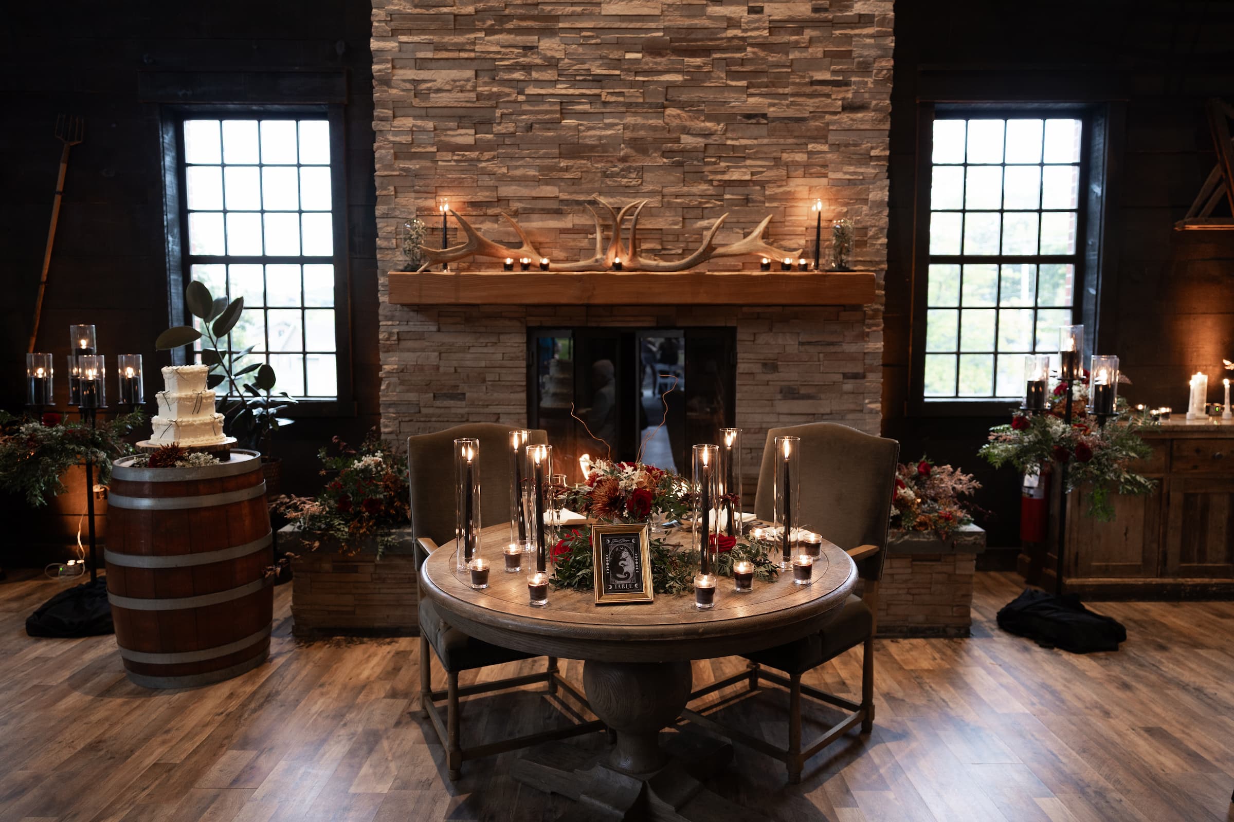 A moody indoor wedding at Diamond Mills Blackbarn with a stone fireplace, dark wood walls, and a round wooden table set with candles, chairs, and floral arrangements. A barrel with a tiered wedding cake stands to the left, and windows with black frames are visible on both sides.