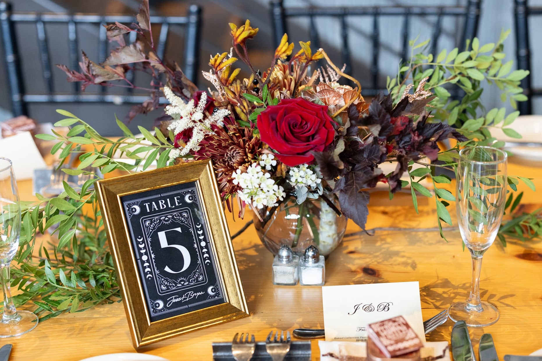 A moody wedding table setting featuring a central floral arrangement with a prominent red rose, green foliage, and a gold-framed table number 5 sign at Diamond Mills Blackbarn. The table is set with white plates, silver cutlery, wine glasses, and a small card with a cake topper photo.