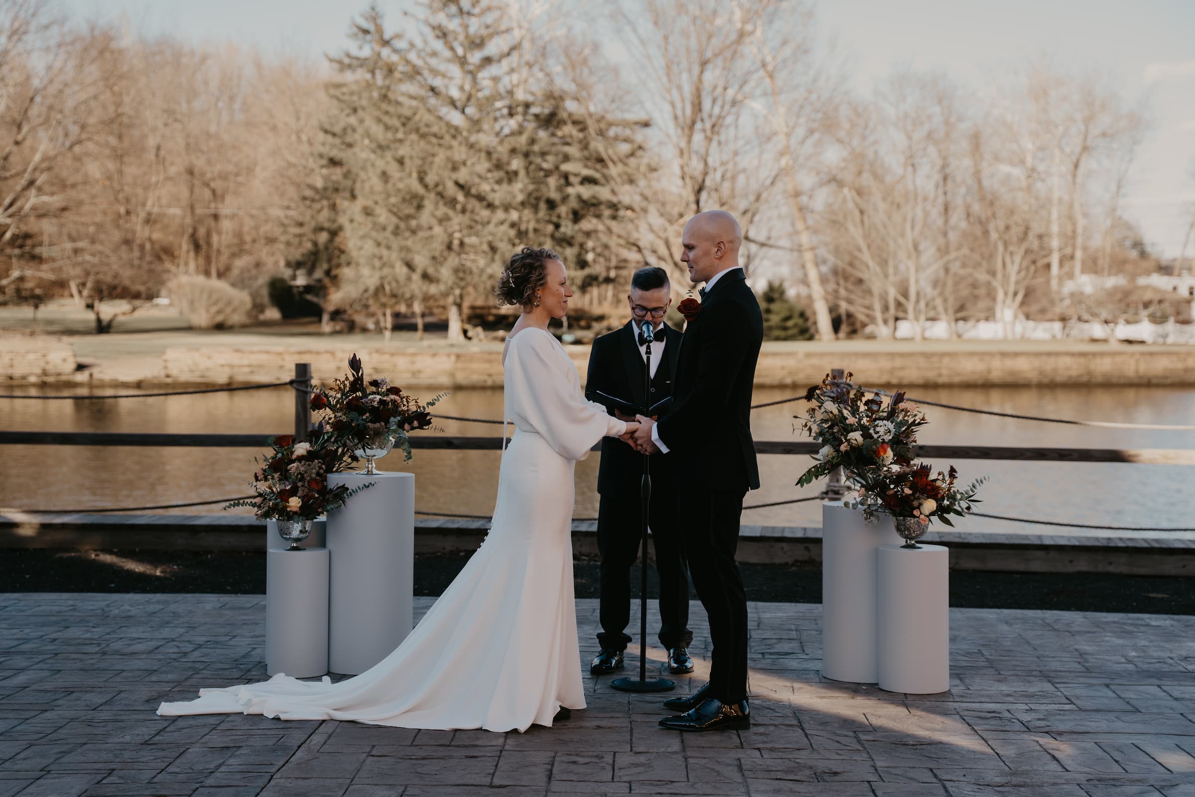 A bride in a long white wedding gown and a groom in a black suit stand facing each other outdoors at the Diamond Mills Boathouse, with a officiating minister in the center and floral arrangements on white pedestals.