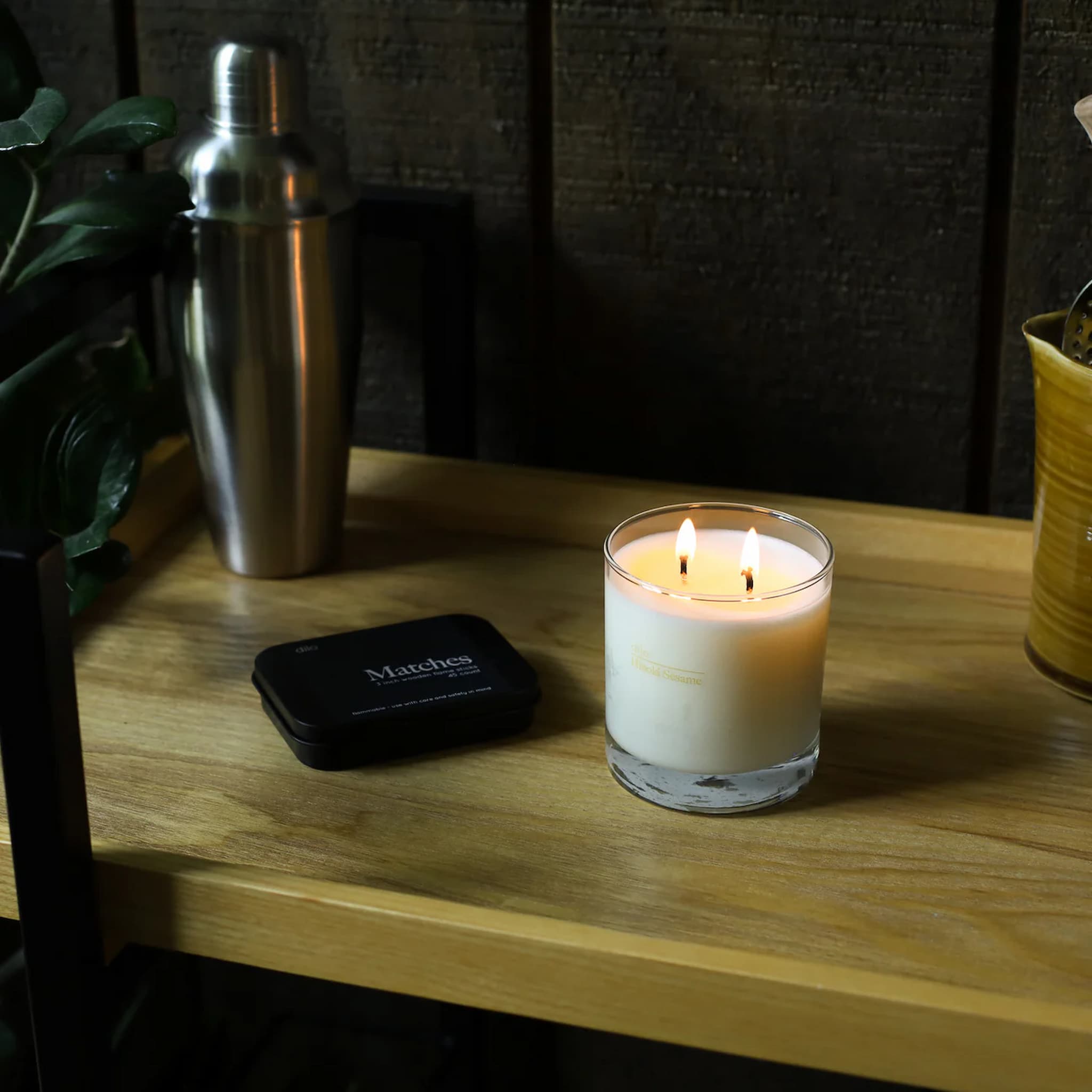 Candle burning on a wooden surface with a cocktail shaker and matches nearby.